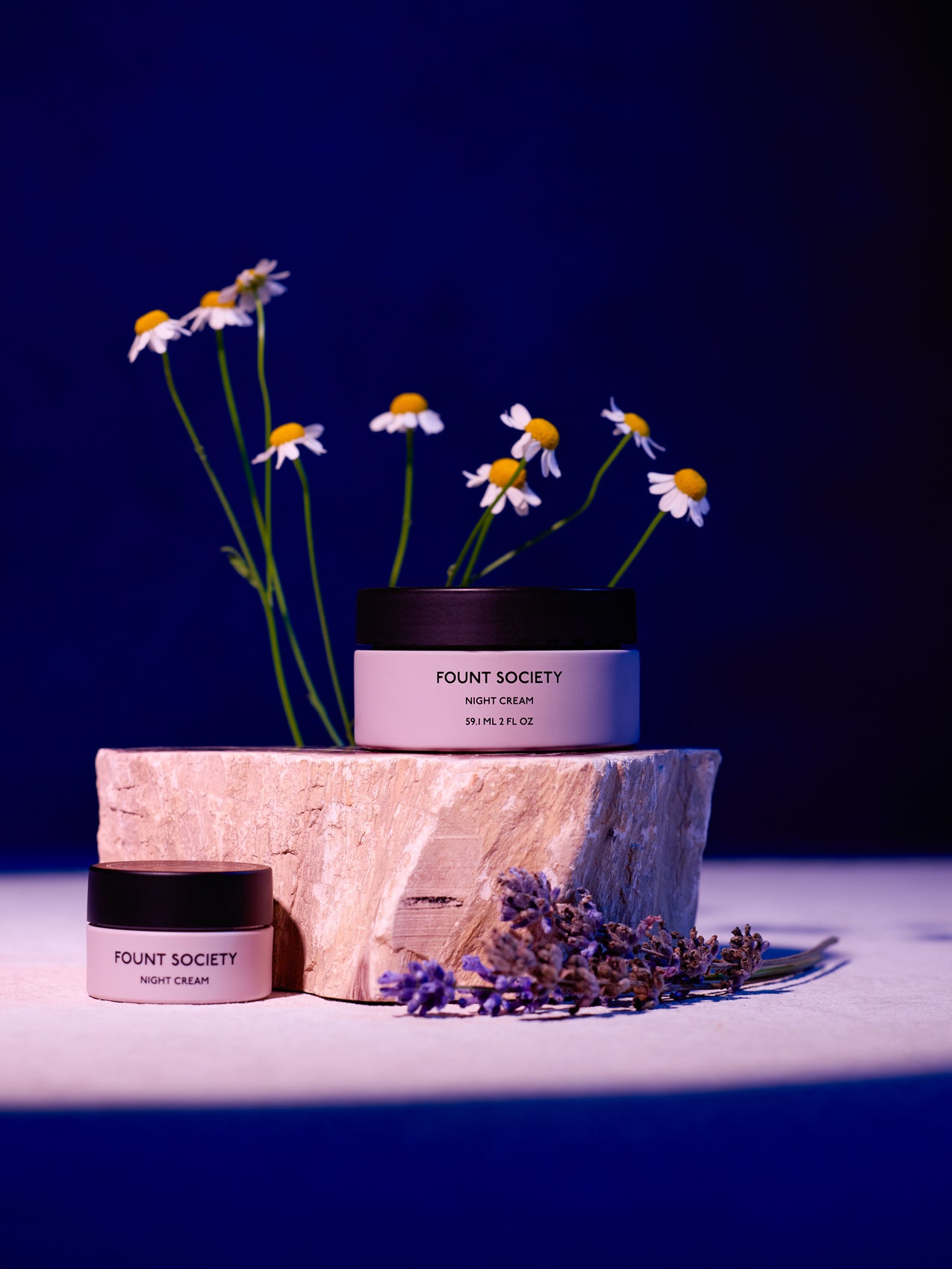 Two jars of Cozy Earth Night Cream sit on a wooden stump with a deep blue backdrop, surrounded by white daisies and purple flowers that enhance the product's natural elegance. 