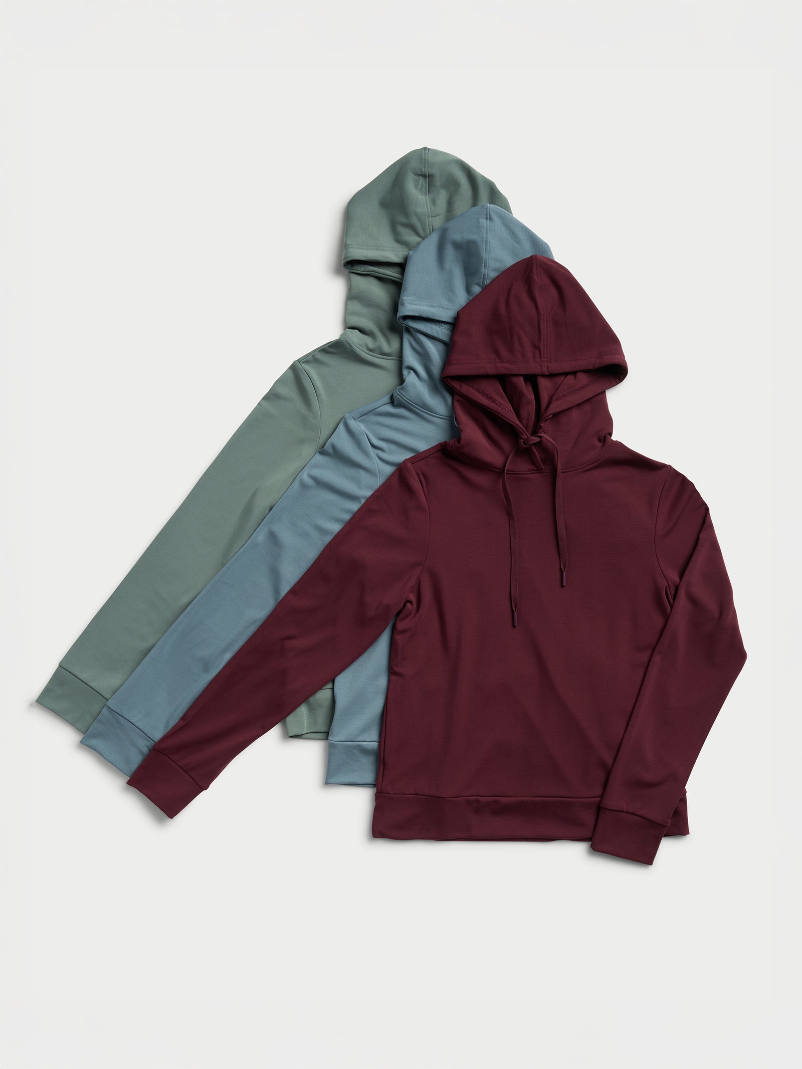 A trio of Women's Ultra-Soft Bamboo Hoodies by Cozy Earth is displayed in a row on a light background. The colors include burgundy, light blue, and green, and each hoodie features a drawstring and long sleeves. 