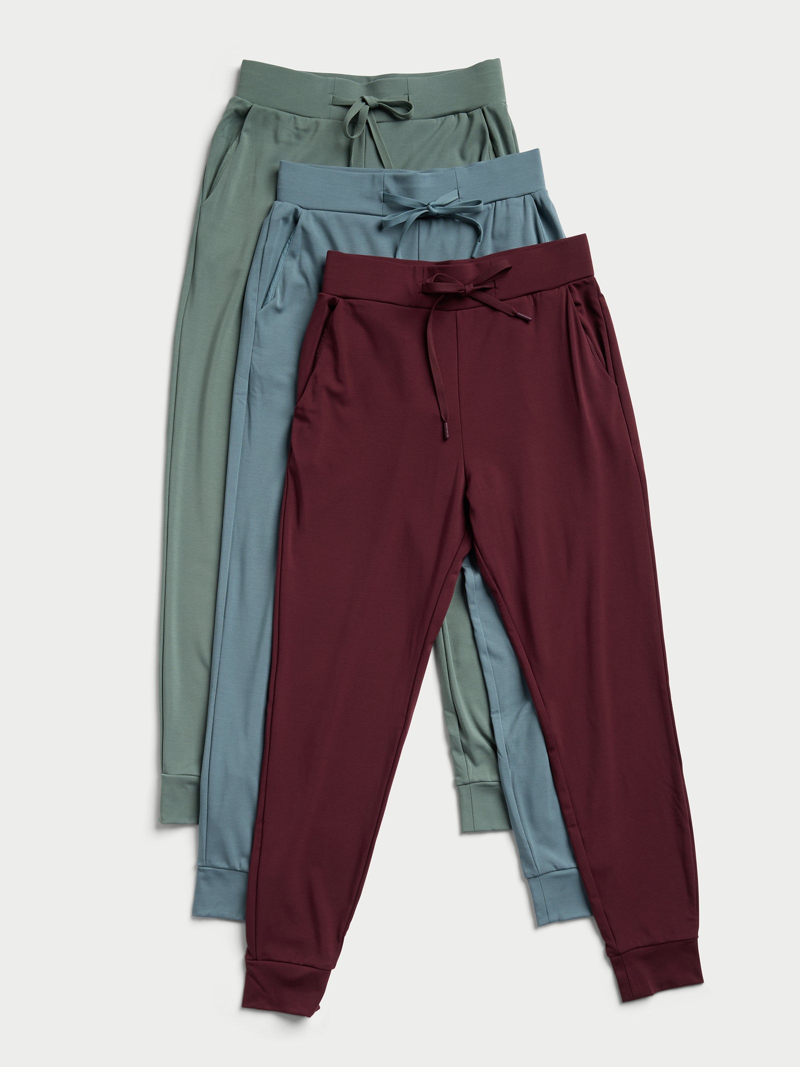 Three pairs of Cozy Earth's Women's Bamboo Jogger Pants TALL in olive green, teal, and maroon are stacked on top of each other. Each pair is designed with an elastic waistband, a drawstring, and cuffed ankles. They are showcased against a light background. 