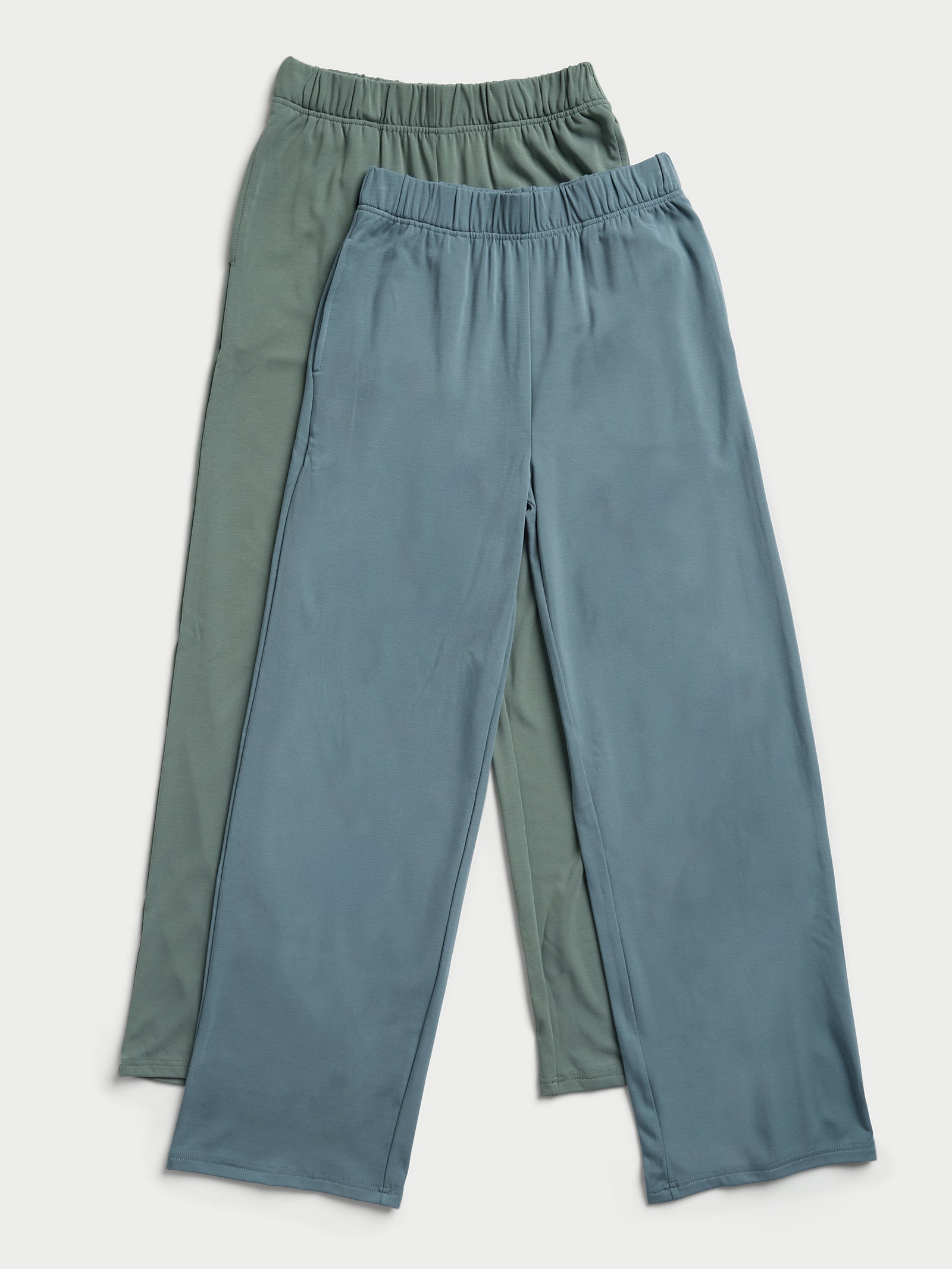 Two pairs of Cozy Earth's Women's Ultra-Soft Bamboo Wide Leg Pull On Pants are overlapping. One pair is in a light sage green color, while the other is in muted teal. Both feature elastic waistbands and straight legs, resting against a light background. 