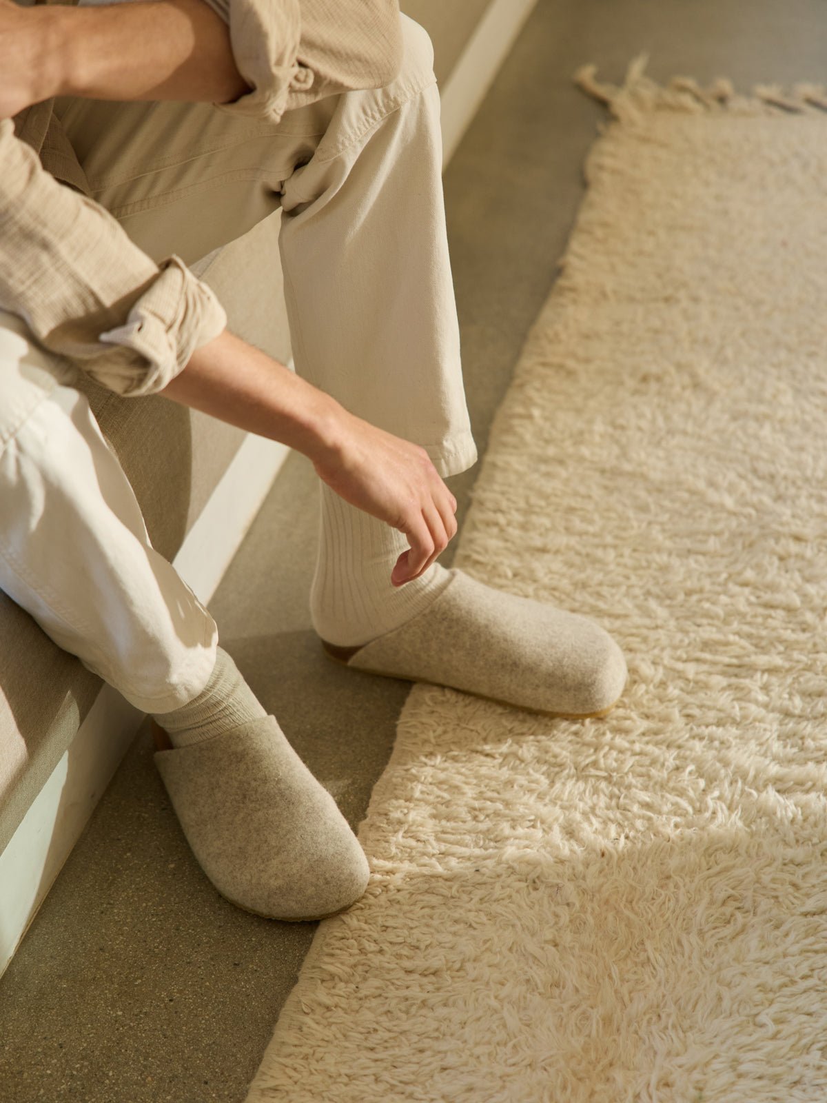 A person dressed in cream-colored loungewear and Cozy Earth's Lakehouse Clog sits on a couch's edge. Their hand rests on their thigh, and a textured cream rug covers the floor while natural light casts soft shadows across the scene. 