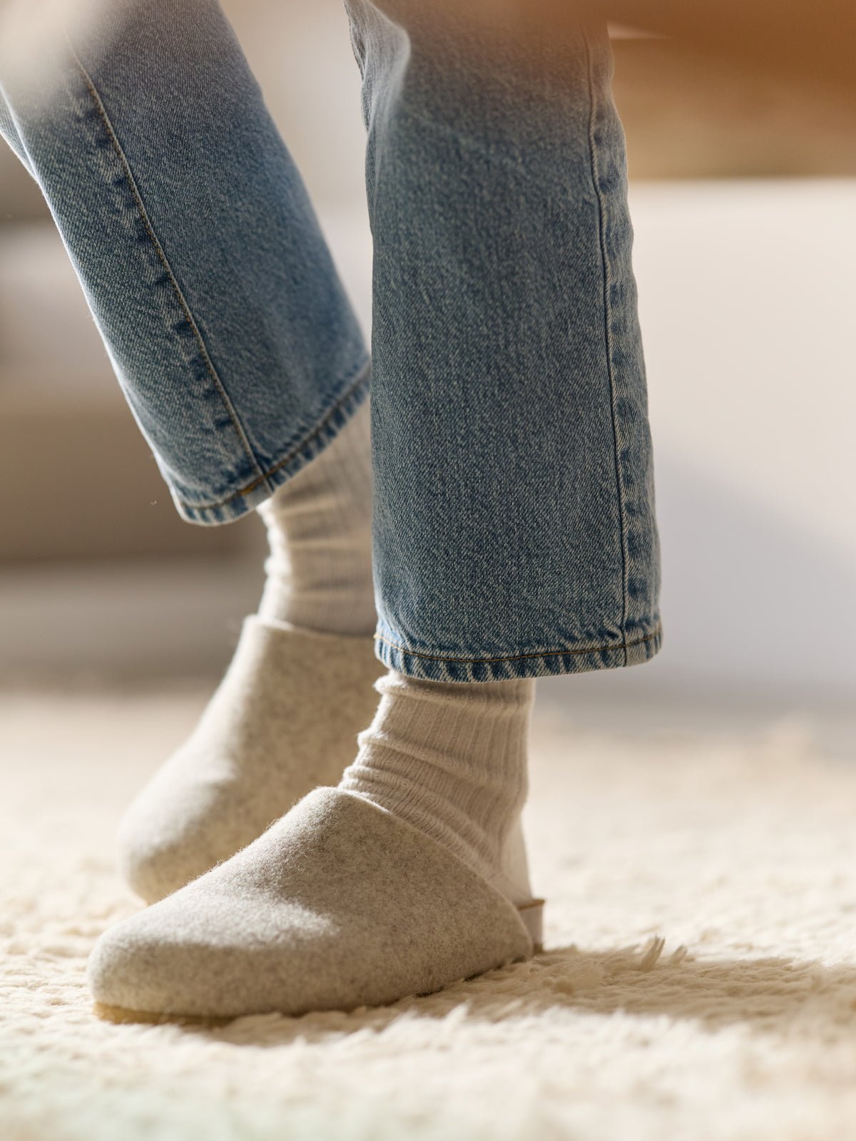 A person in blue jeans and white socks stands on a cream-colored textured rug, wearing Lakehouse Clogs by Cozy Earth. 