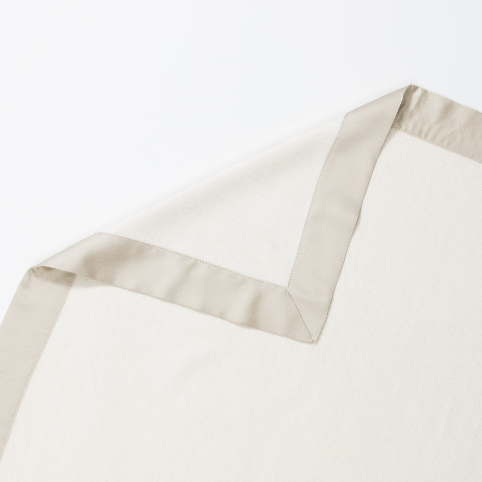 The Bamboo Blanket by Cozy Earth, featuring an off-white hue and a light beige satin trim, is elegantly laid out on a white surface. One corner of the blanket is folded over, revealing that the underside shares the same color and texture as the top. 