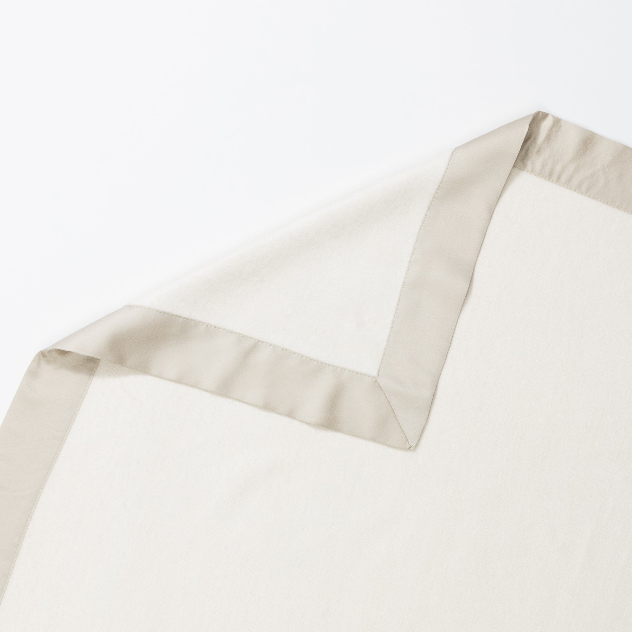 The Bamboo Blanket by Cozy Earth, featuring an off-white hue and a light beige satin trim, is elegantly laid out on a white surface. One corner of the blanket is folded over, revealing that the underside shares the same color and texture as the top. |Color:Oat/Driftwood