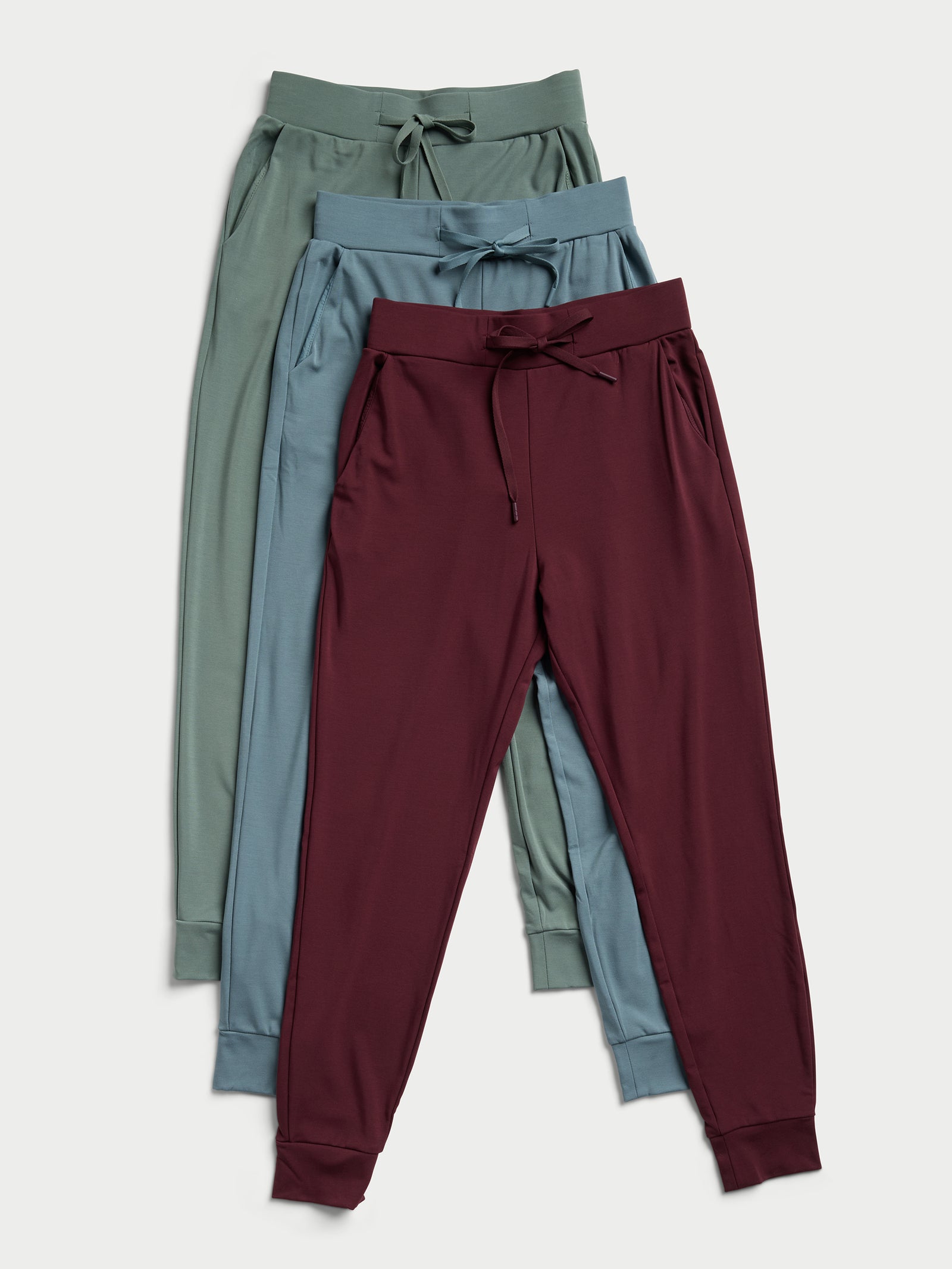 Three pairs of the Women's Bamboo Jogger Pant by Cozy Earth, in green, blue, and maroon, are placed overlapping each other. Each pair showcases a drawstring waist and cuffed ankles against a light background. 