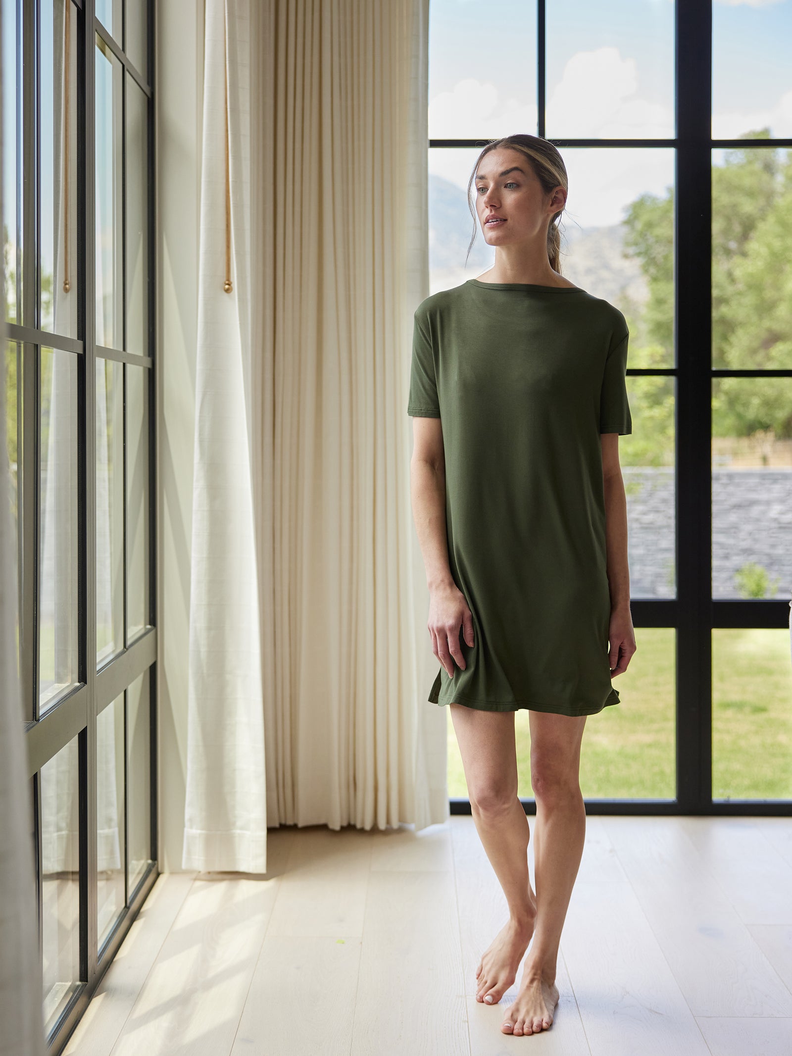 A woman stands barefoot by tall windows in a bright room. She is wearing the Cozy Earth Women's Bamboo Stretch Knit Sleep Dress, a simple, short-sleeved green dress. The floor is pale wood, and light curtains frame the windows. Greenery and hills can be seen outside. The woman appears relaxed and contemplative. 