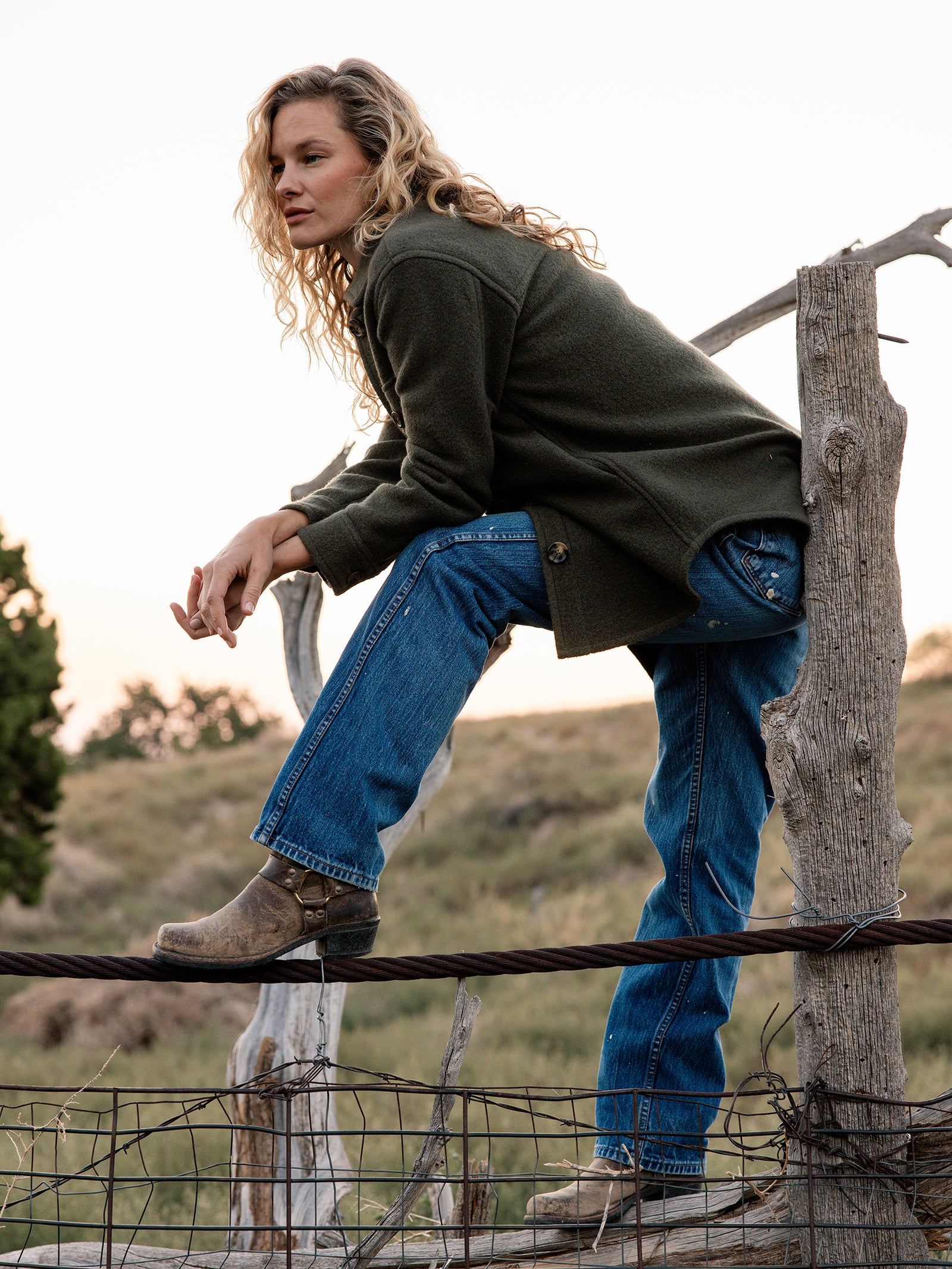 A person with long curly hair, wearing a Cozy Earth Women's Boucle Shacket, blue jeans, and brown boots, is sitting on a rustic wooden fence in a rural outdoor setting. The person appears to be gazing into the distance as the sun sets. 