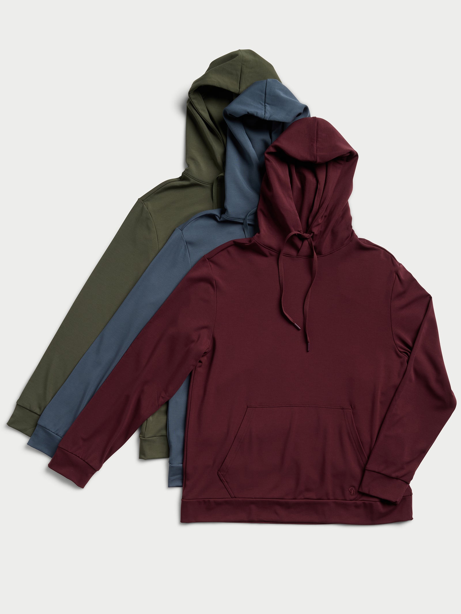 Three Men's Ultra-Soft Bamboo Hoodies by Cozy Earth are arranged side by side on a neutral background. The pullovers come in earthy tones: olive green, deep teal, and maroon. Each pullover features a front pocket and drawstrings. 