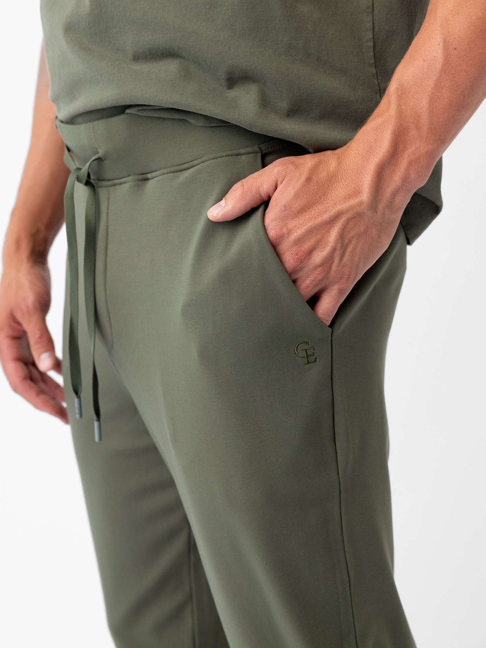 A person is wearing Cozy Earth's Men's Ultra-Soft Bamboo Jogger Pant in olive green, which features a drawstring waist and a relaxed fit. Their right hand is placed inside the pant pocket, which has a small embroidered logo. The person is also sporting a matching olive green top. 