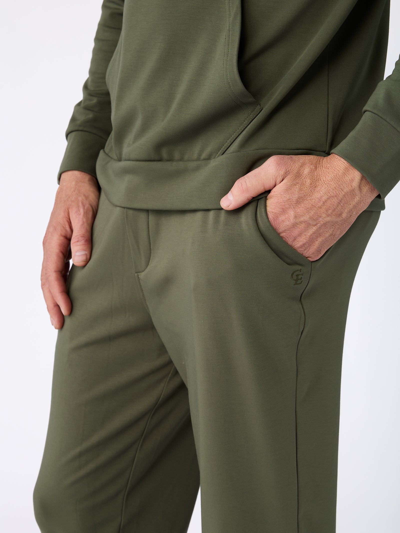 A person wearing the Cozy Earth Men's Ultra-Soft Bamboo Pullover Crew in olive green is seen with their right hand in their pocket, paired with joggers. The outfit is crafted from a soft, comfortable fabric. The background is a simple, plain white. 