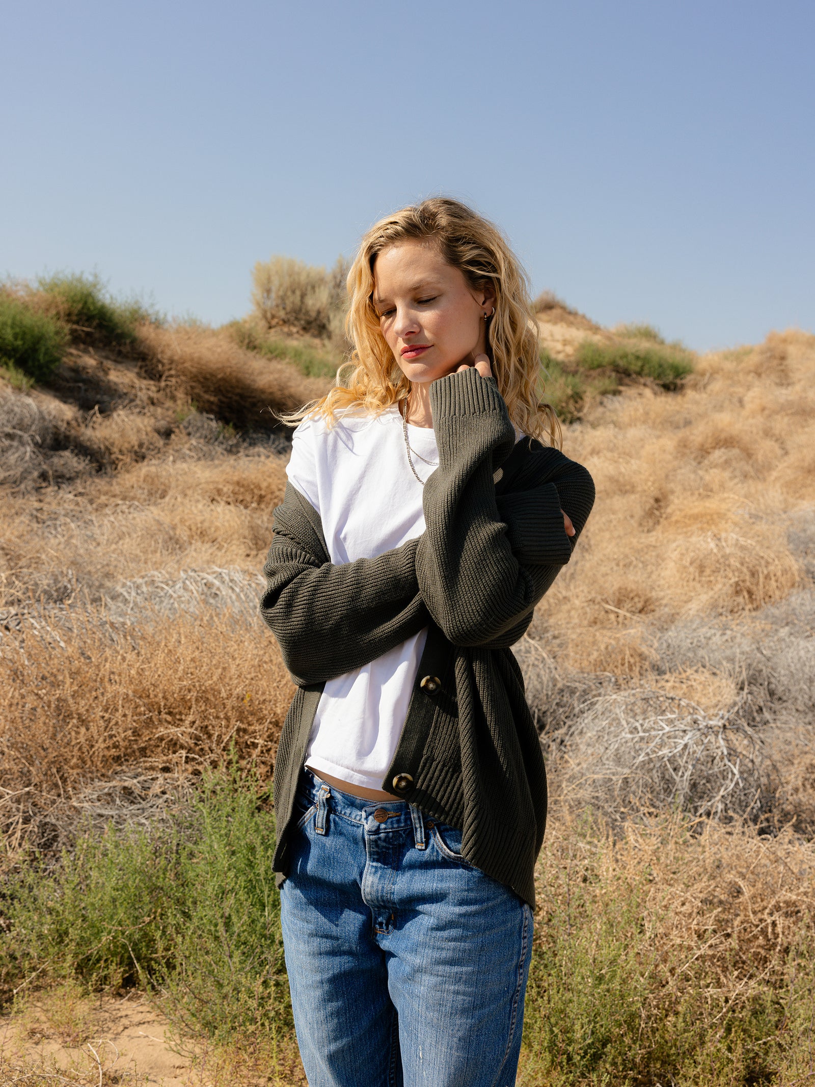 A person with long, wavy blonde hair stands outdoors in a dry, grassy landscape. They are wearing a white T-shirt paired with the Cozy Earth Oversized Classic Cardigan and blue jeans, arms crossed and eyes closed, under a clear blue sky. 