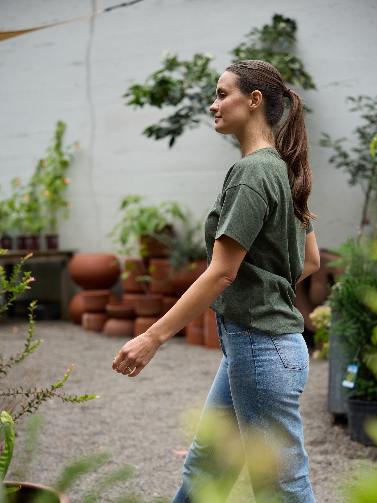 A woman with a ponytail and wearing the Cozy Earth Women's All Day Tee in green is walking through an outdoor garden center. She is surrounded by potted plants and large terracotta pots, appearing calm and focused as she navigates the space. The background features a concrete wall. 