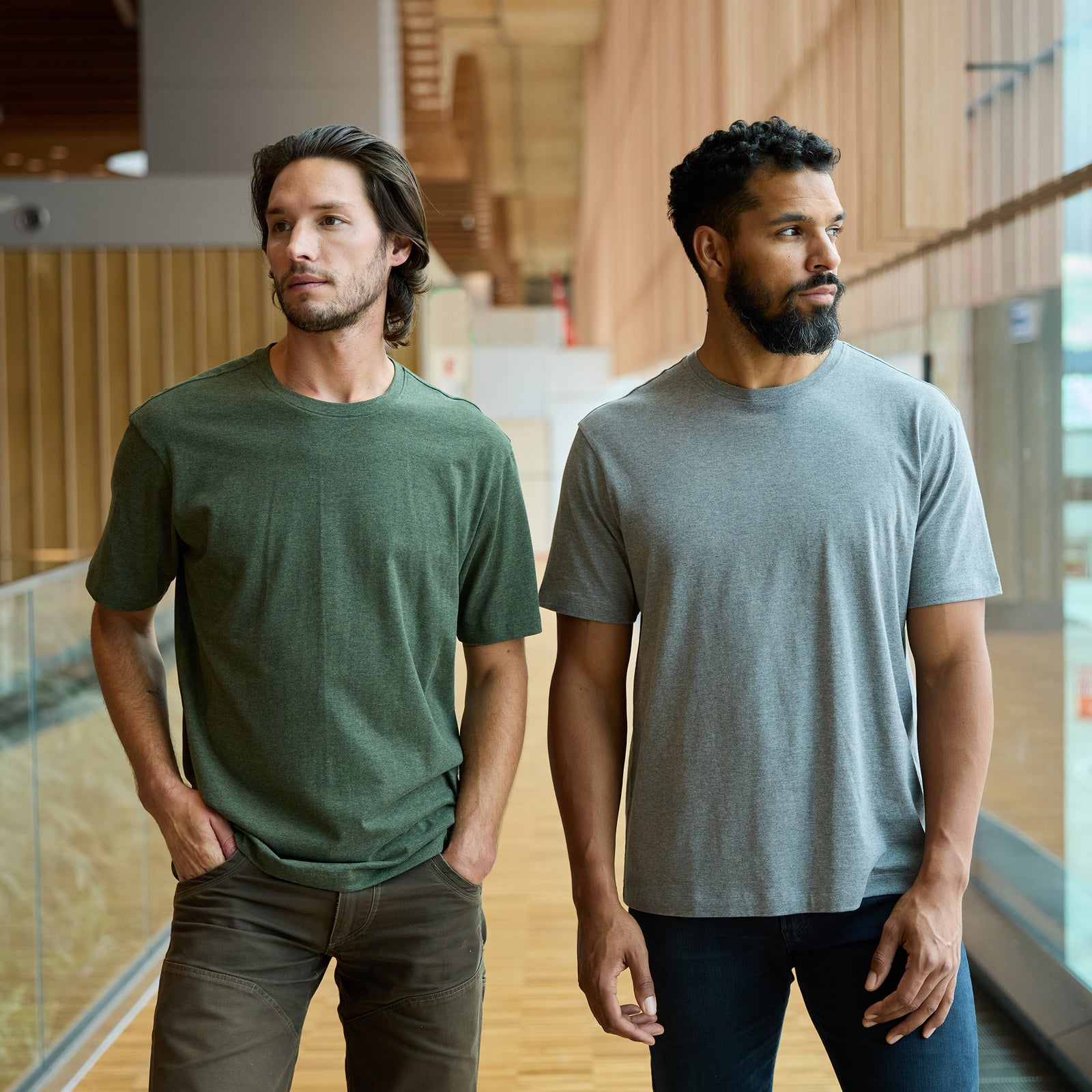 Two men stand side by side in a modern indoor setting with wooden walls and glass railings. The man on the left wears a green Men's All Day Tee by Cozy Earth paired with brown pants, while the man on the right wears a grey Men's All Day Tee by Cozy Earth combined with dark blue jeans. Both look off into the distance. 