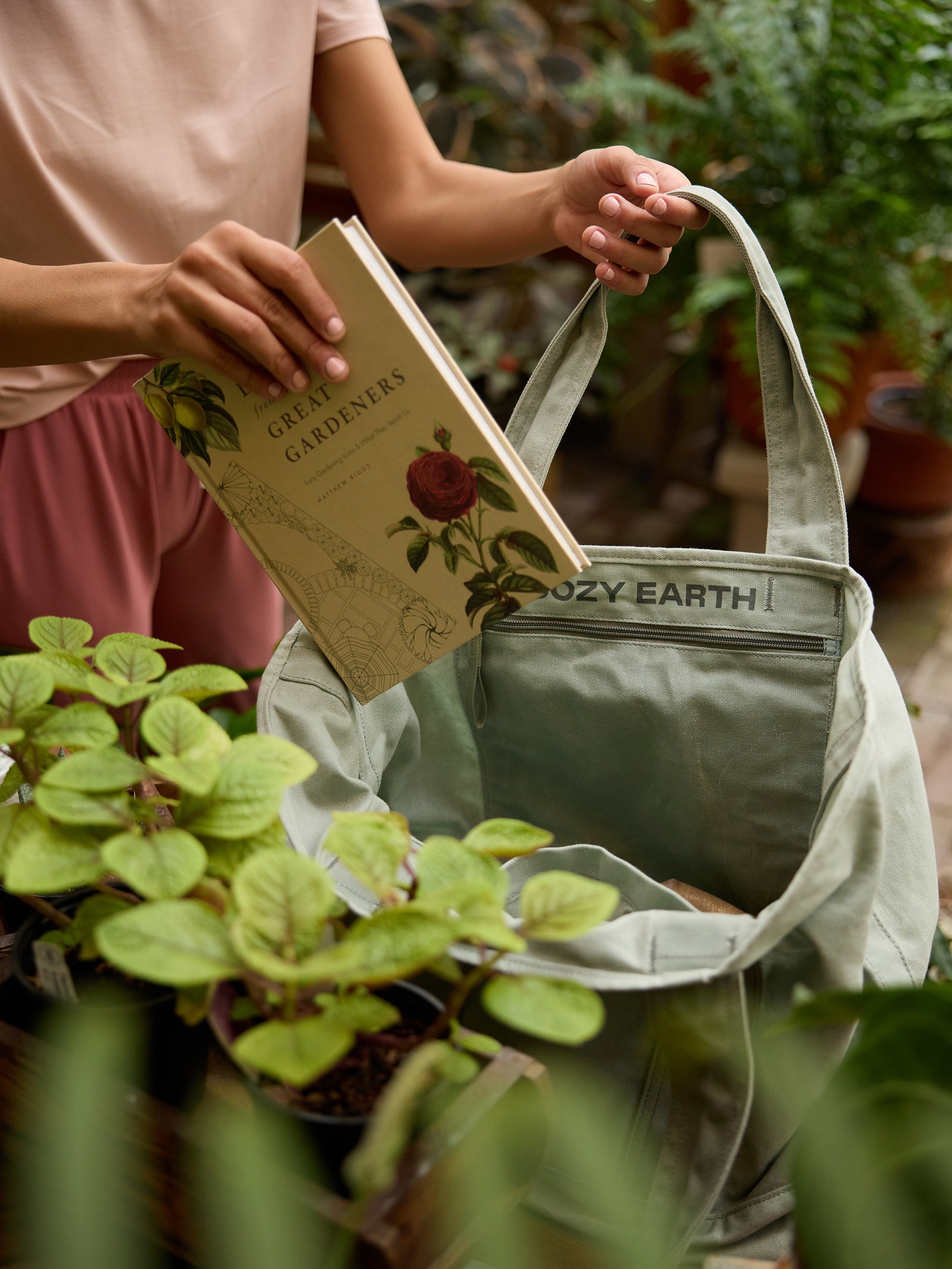 A person in a pink outfit is placing a book titled "Green Gardeners" into a green Waxed Canvas Tote labeled "Cozy Earth." The scene is set in a lush garden with various potted plants surrounding them. 