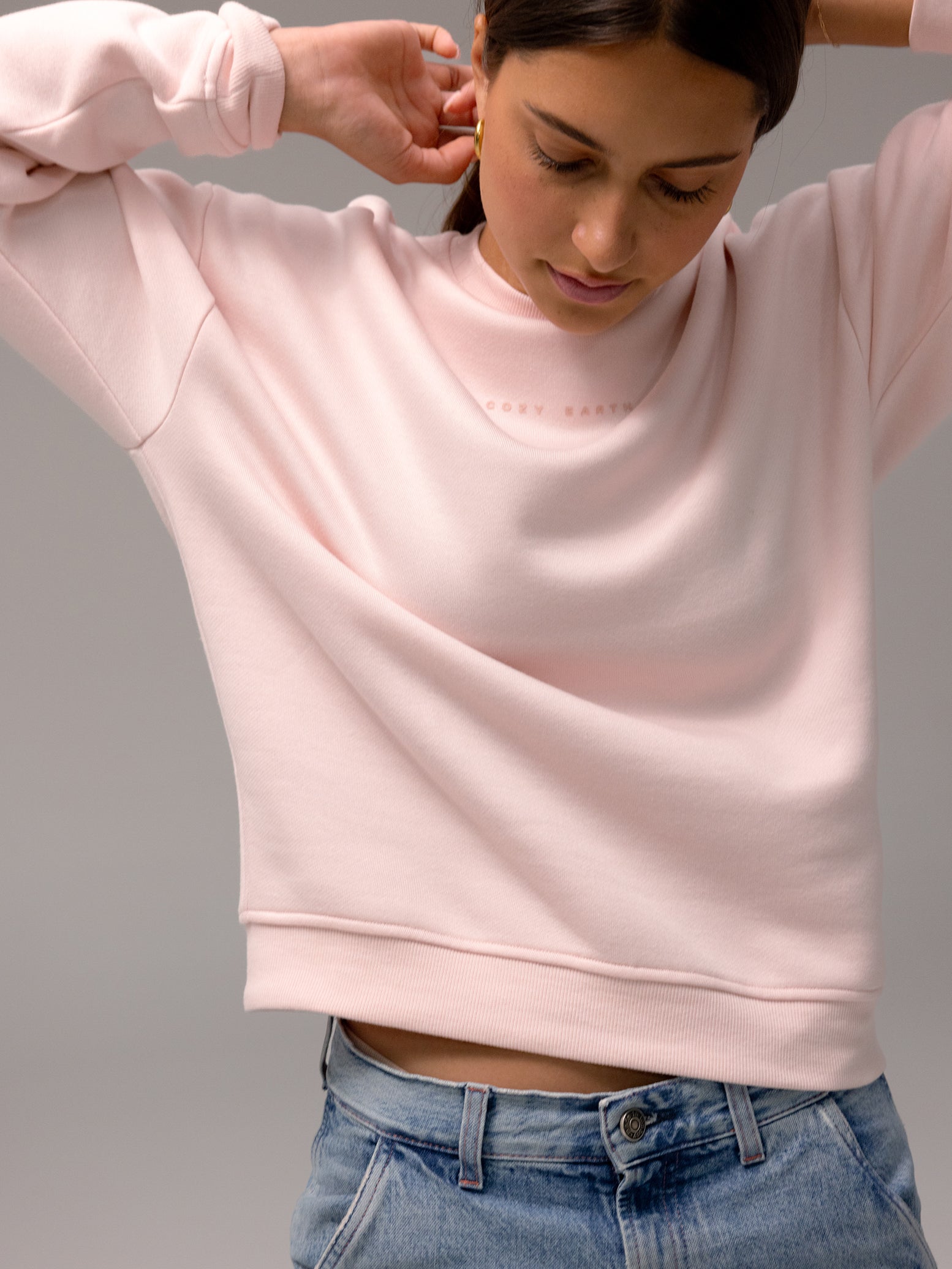 A woman wearing the light pink Women's CityScape Crewneck by Cozy Earth and blue jeans leans forward, looking down, against a plain gray background. 