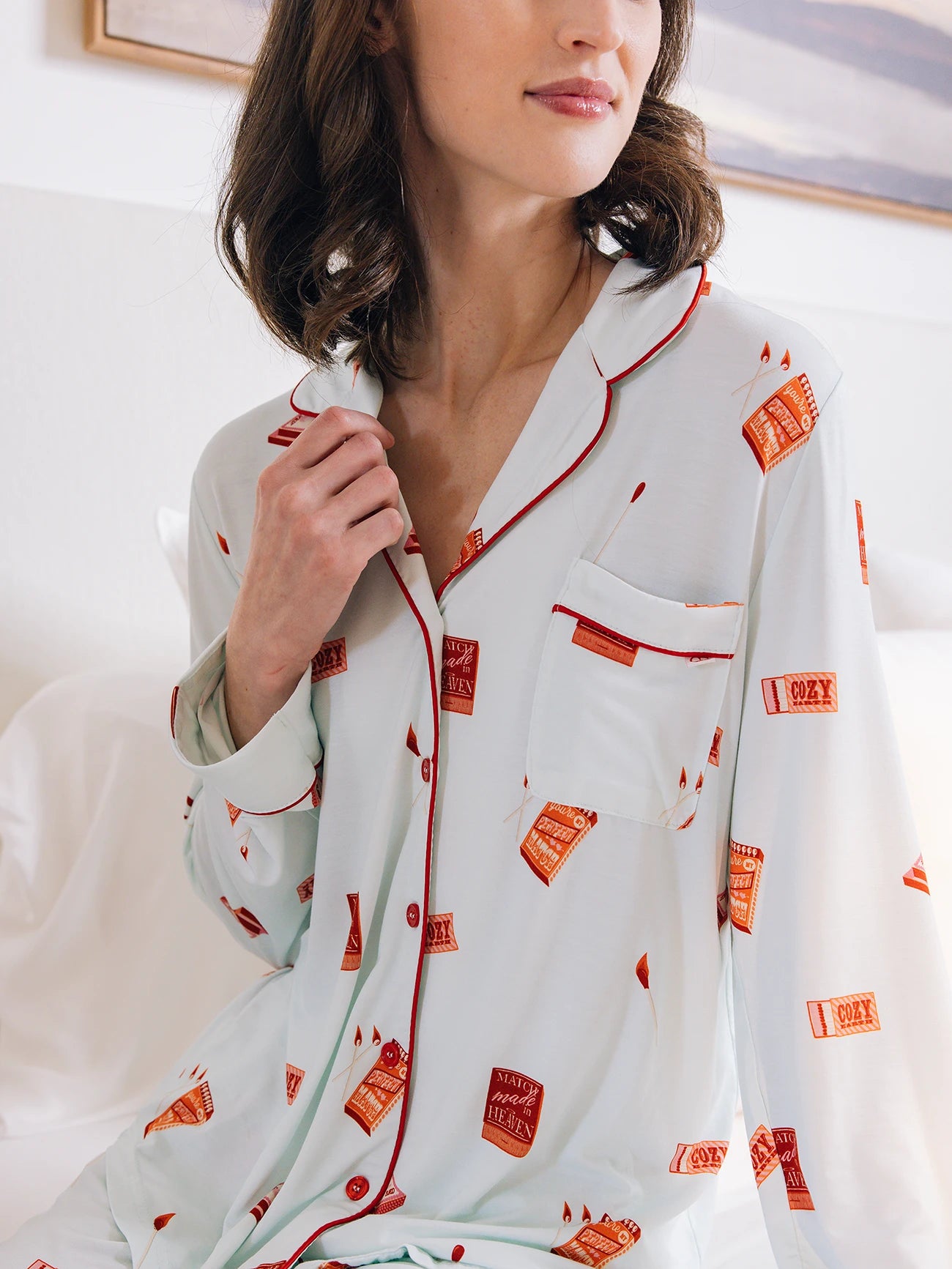 A person is sitting on a bed wearing Cozy Earth Women's Long Sleeve Bamboo Pajama Top in Stretch-Knit, featuring a relaxed fit, white fabric with red piping, and prints of red and orange book covers. The pajamas have a chest pocket and the person is touching the collar. 