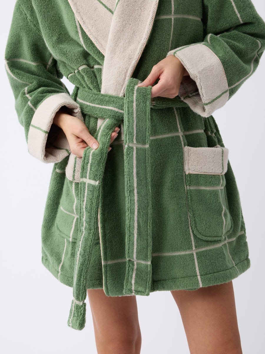 A person fastens the belt of a Cozy Earth Windowpane Resort Robe. The green robe features beige trim, white checkered patterns, and two front pockets. Only their lower body and hands are visible against a plain background. 