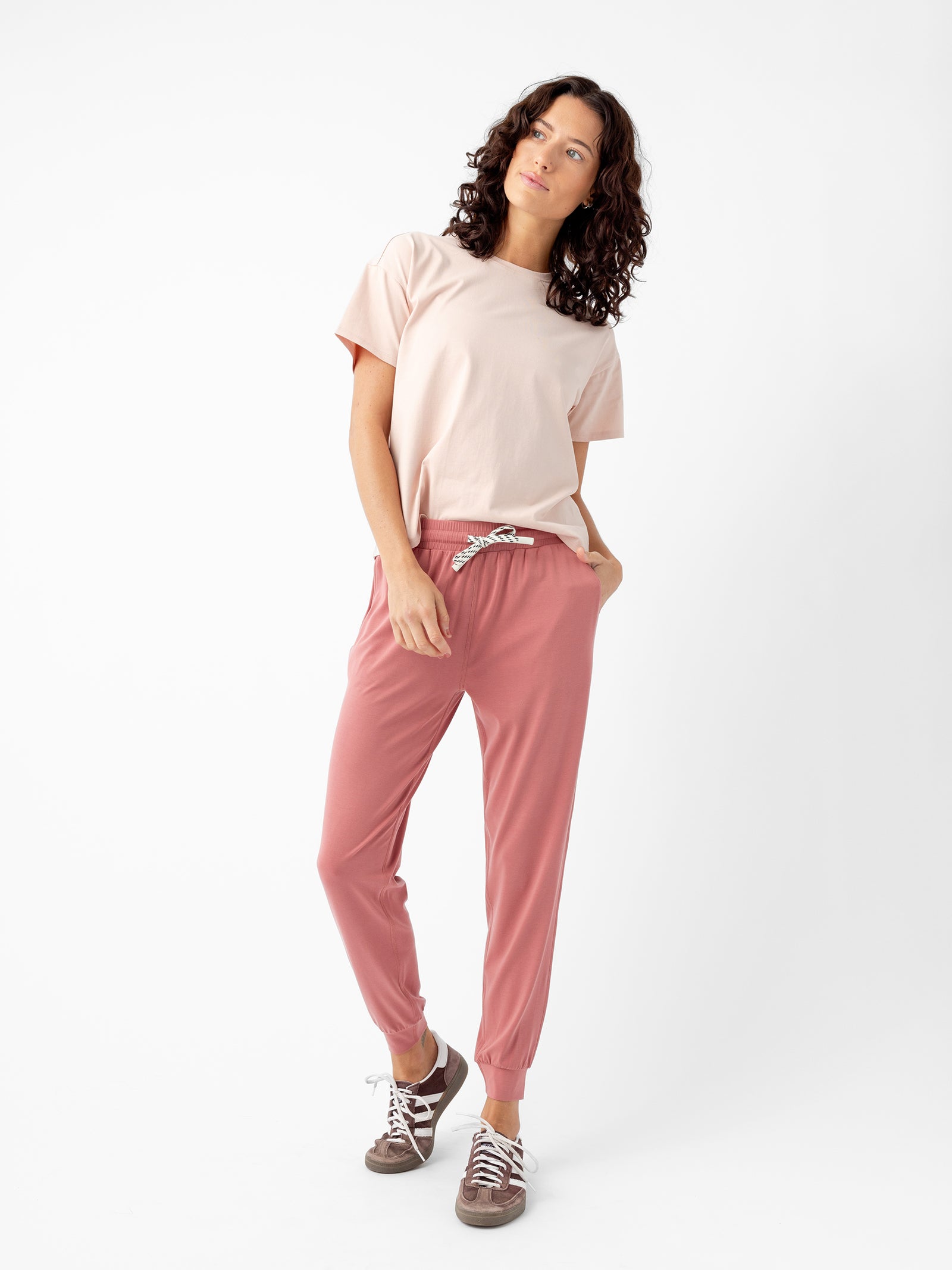 A person with curly hair is standing against a plain white background, wearing a light pink t-shirt and Women's Studio Jogger pants by Cozy Earth, tied at the waist with a white drawstring. They are also wearing brown sneakers and have their hands in their pockets, gazing slightly upwards. 