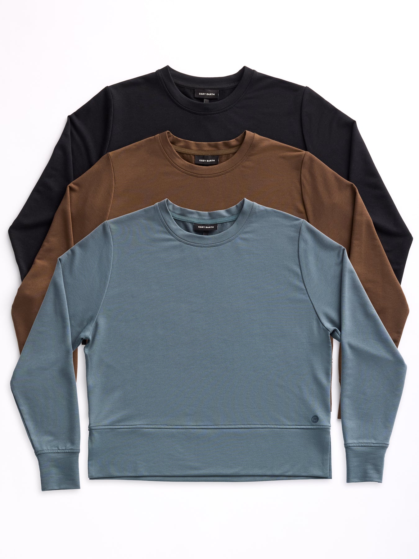 Three Women's StudioLite Crewneck sweaters from Cozy Earth are neatly stacked against a plain white background. The bottom sweater is blue, the middle one is brown, and the top one is black. 