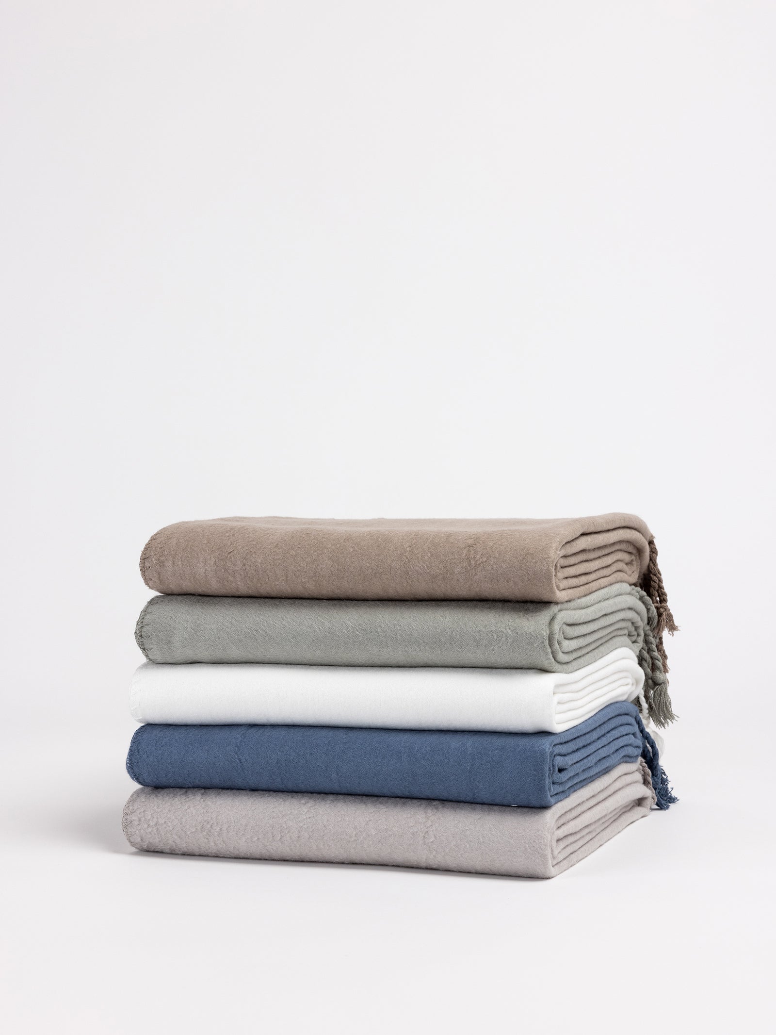 A stack of four Cozy Earth Bamboo Tassel Throws in different colors sits against a plain white background. From top to bottom, the throws are brown, green, white, and blue in color. The edges feature a subtle fringe detailing. 