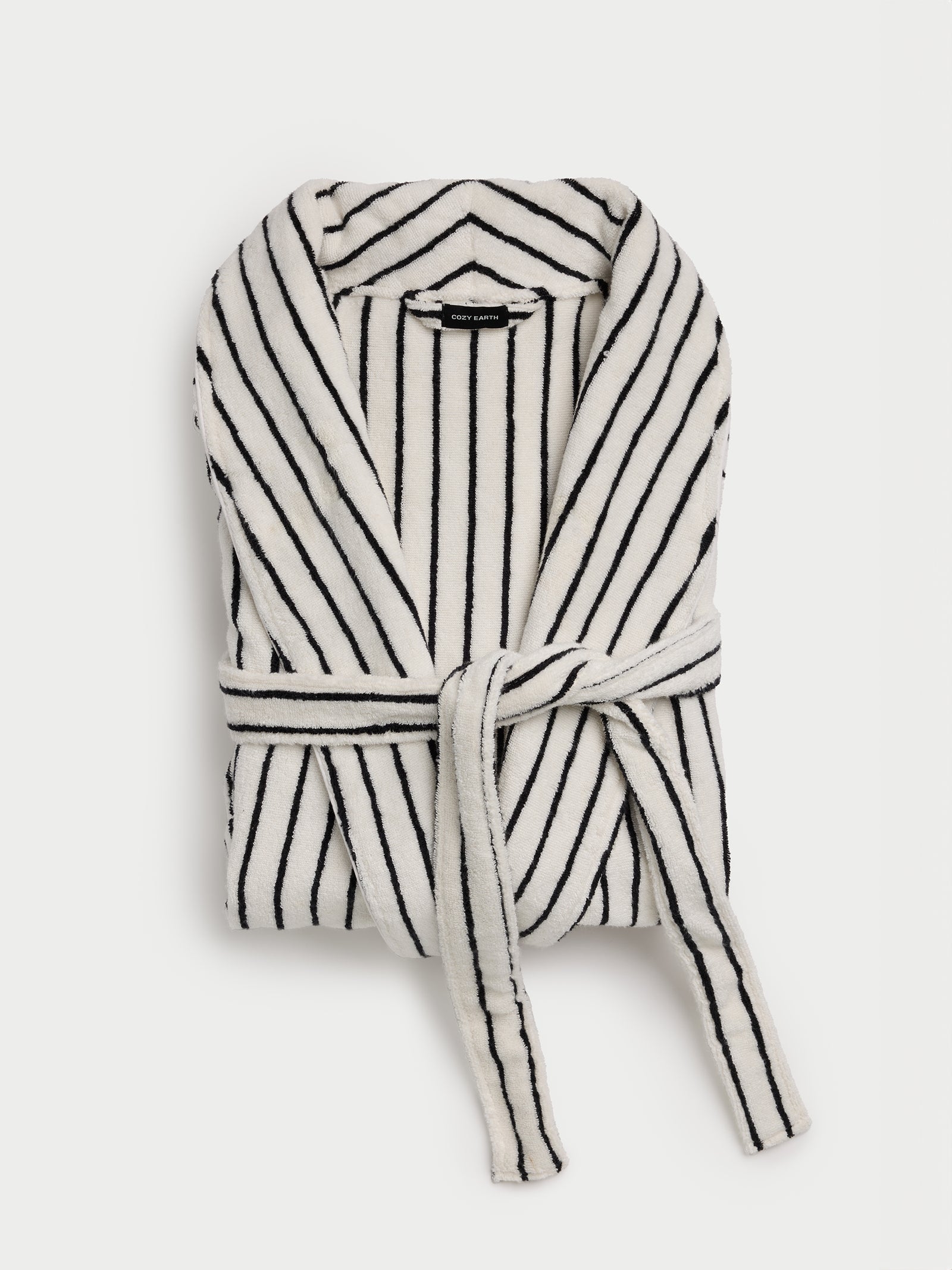 A Luxe Bath Robe from Cozy Earth, featuring a folded white design with black vertical stripes and a matching tie belt, rests on a plain light background. 