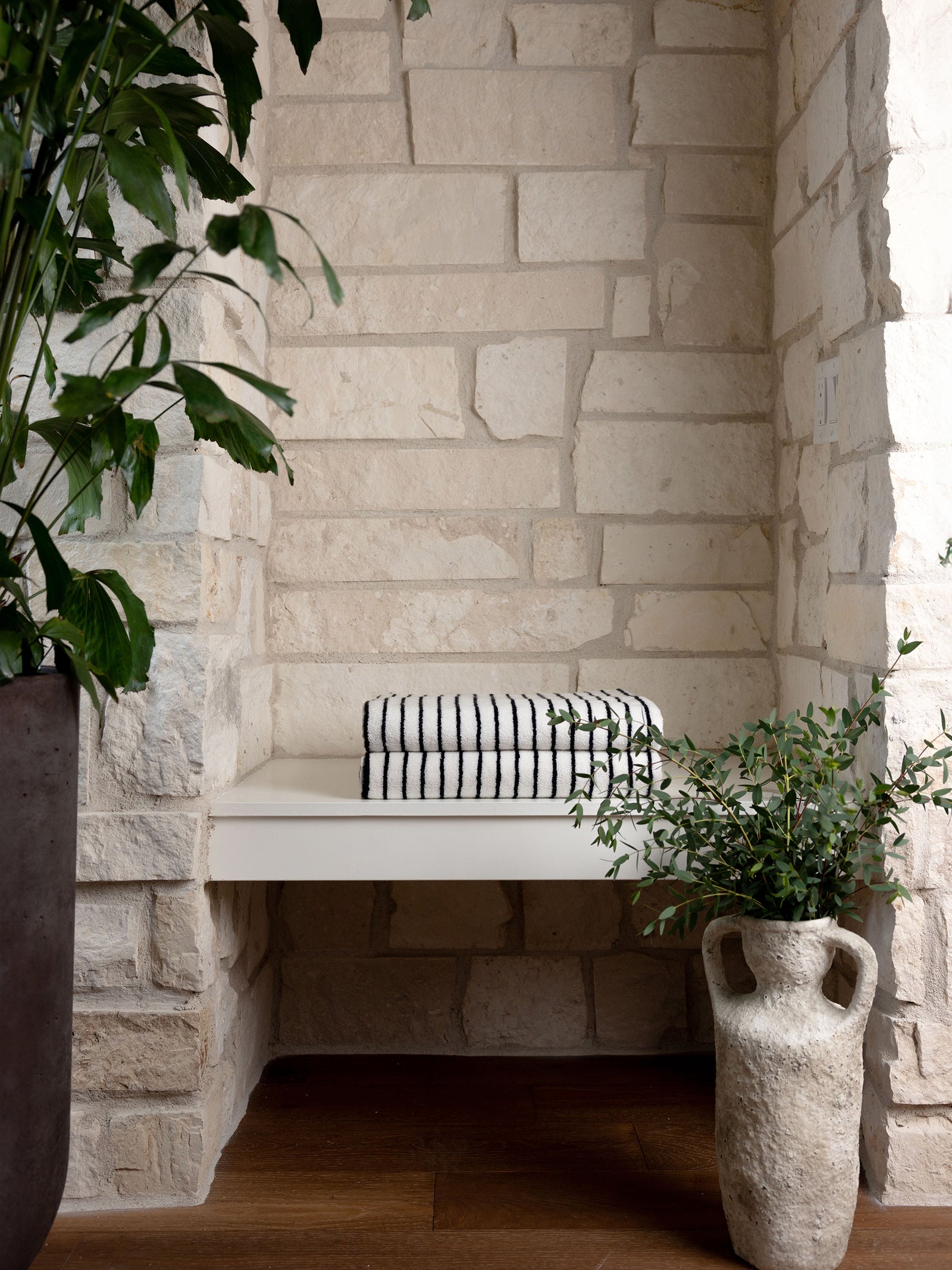 A cozy nook with a built-in white bench against a textured stone wall, featuring Luxe Bath Towels by Cozy Earth in a black and white stripe pattern. The setting is accentuated by lush green plants in pots on the wooden floor, creating a serene and inviting atmosphere. 