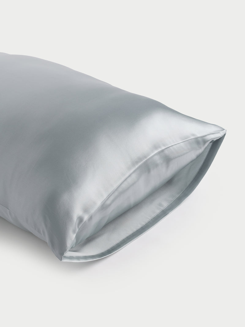 A shiny, light blue Silk Pillowcase by Cozy Earth with a smooth texture is displayed against a plain background. The open end is visible, highlighting the fabric's glossy finish.