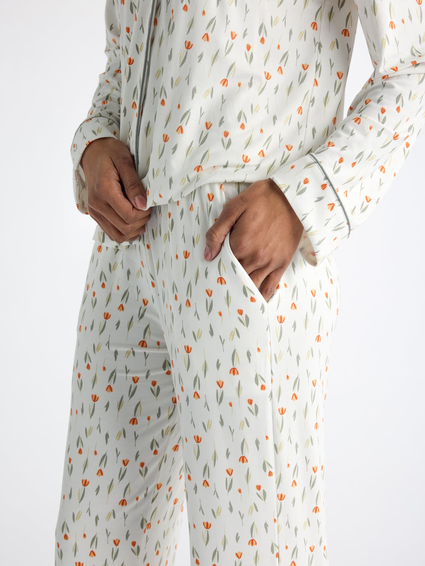 A person in Cozy Earth's Women's Bamboo Stretch-Knit Long Sleeve Pajama Set, featuring an orange and green floral pattern, stands with one hand in the pocket and the other adjusting the fabric against a plain white background. 