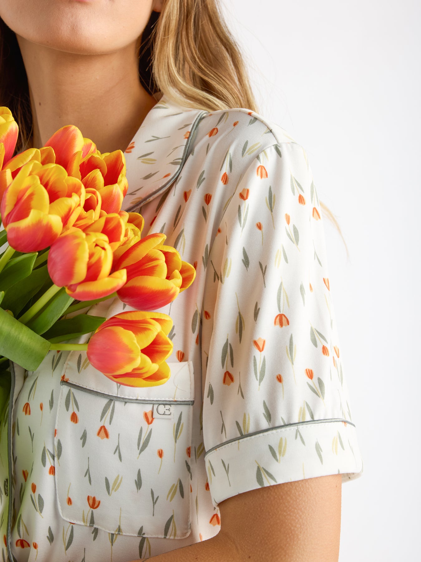 A woman holds a bouquet of orange and yellow tulips while wearing a HIDE Women's Bamboo Stretch-Knit Short Sleeve Pajama Top with a tulip pattern. The image is cropped above her mouth, highlighting the colorful flowers and patterned shirt. 