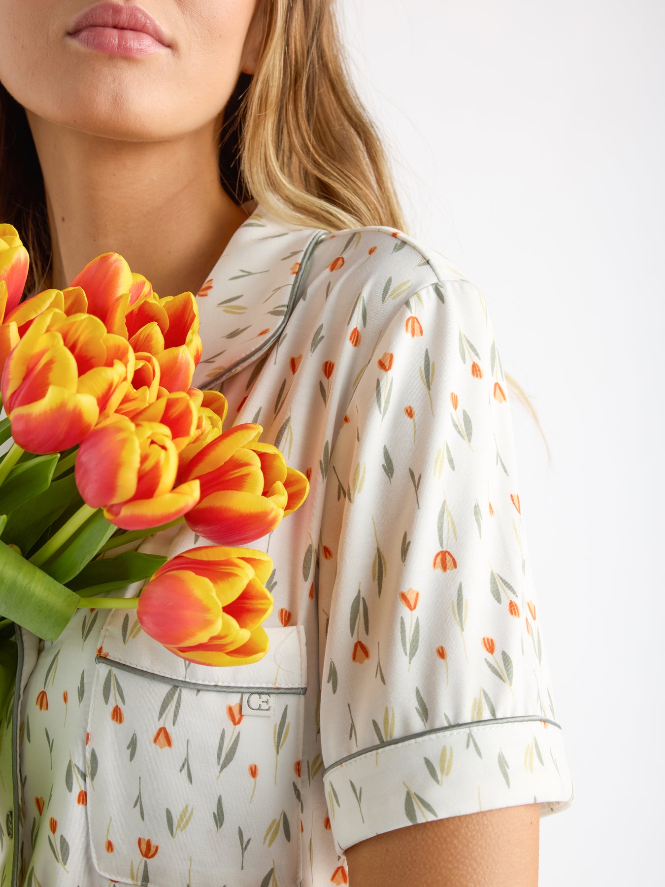 A person in a Women's Bamboo Stretch-Knit Short Sleeve Pajama Set by Cozy Earth holds a bouquet of orange and yellow tulips. The pajama set features small orange and green flower designs, with long hair flowing down the partially visible face. 