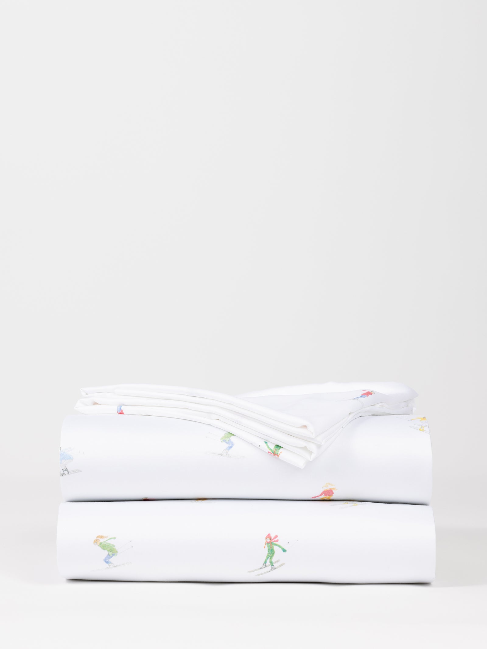 A neatly folded stack of Cozy Earth's Bamboo Sheet Set, featuring a plain white background adorned with small colorful illustrations of people in action poses like surfing and playing sports. 