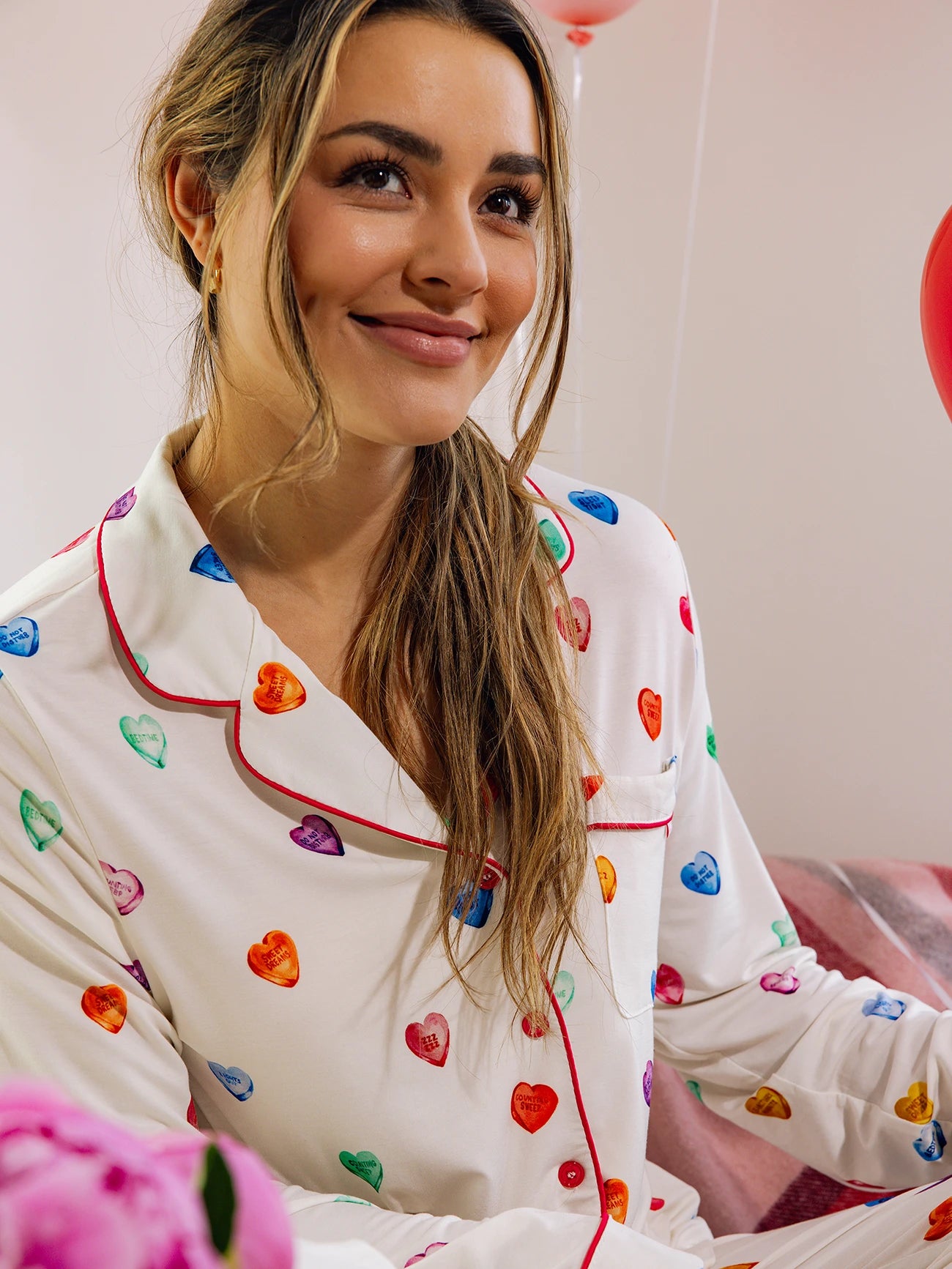 A person with long hair smiles in the brightly lit room, wearing a Cozy Earth Women's Long Sleeve Bamboo Pajama Top in Stretch-Knit, adorned with colorful heart patterns. A red balloon is partially visible nearby, and a pink object sits in the foreground. 