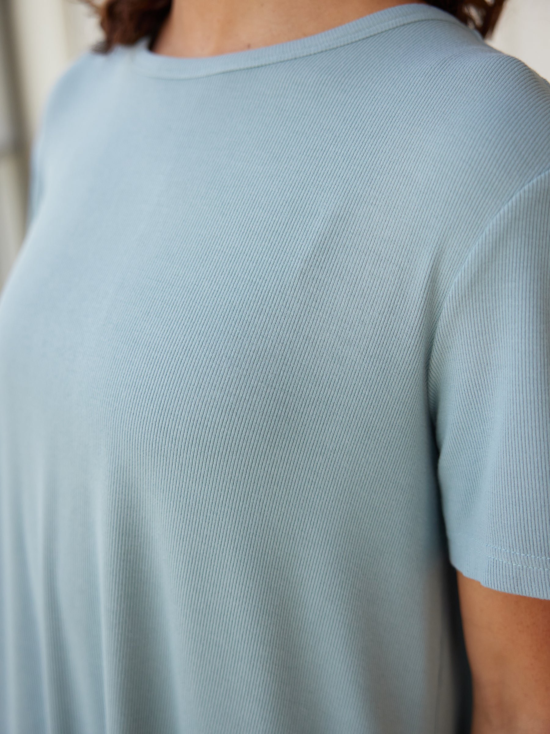 Close-up of a person wearing the Women’s Bamboo Rib-Knit Boyfriend Sleep Dress in light blue by Cozy Earth. The image focuses on the texture and fabric of the dress, showcasing detailed ribbing and soft material. The person's right shoulder and neck area are partially visible. 