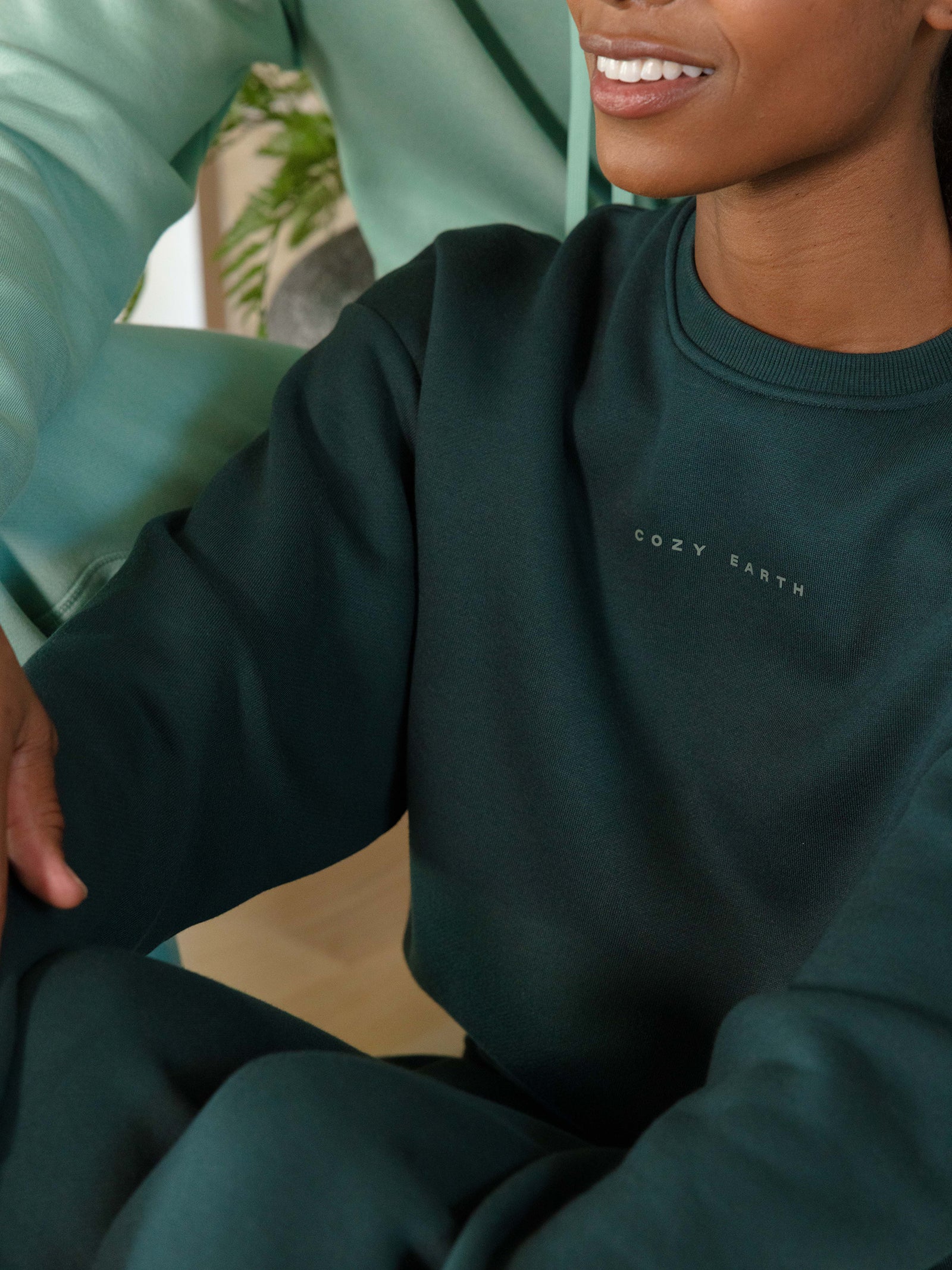 A person wearing a dark green Women's CityScape Crewneck from Cozy Earth is sitting indoors. The image focuses on the upper body and part of the arm, with a soft smile visible. A blurred plant is in the background. 