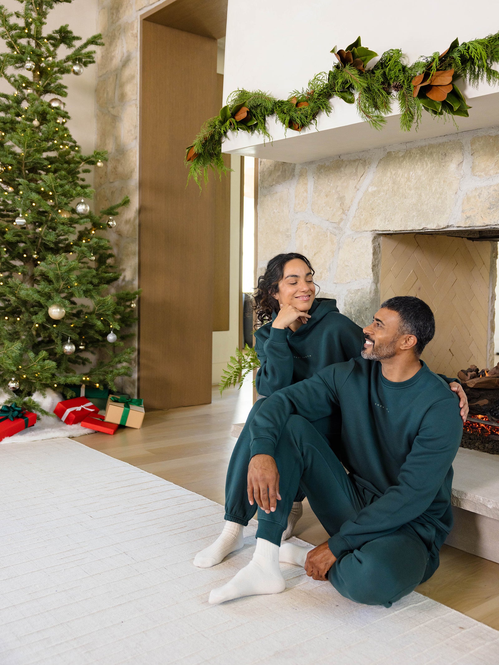 Dressed in matching Men's CityScape Crewnecks by Cozy Earth, a couple sits by the fireplace with warm smiles. A beautifully decorated Christmas tree and an array of gifts are visible in the background, while the mantel boasts lush greenery, creating a cozy and festive holiday atmosphere. 