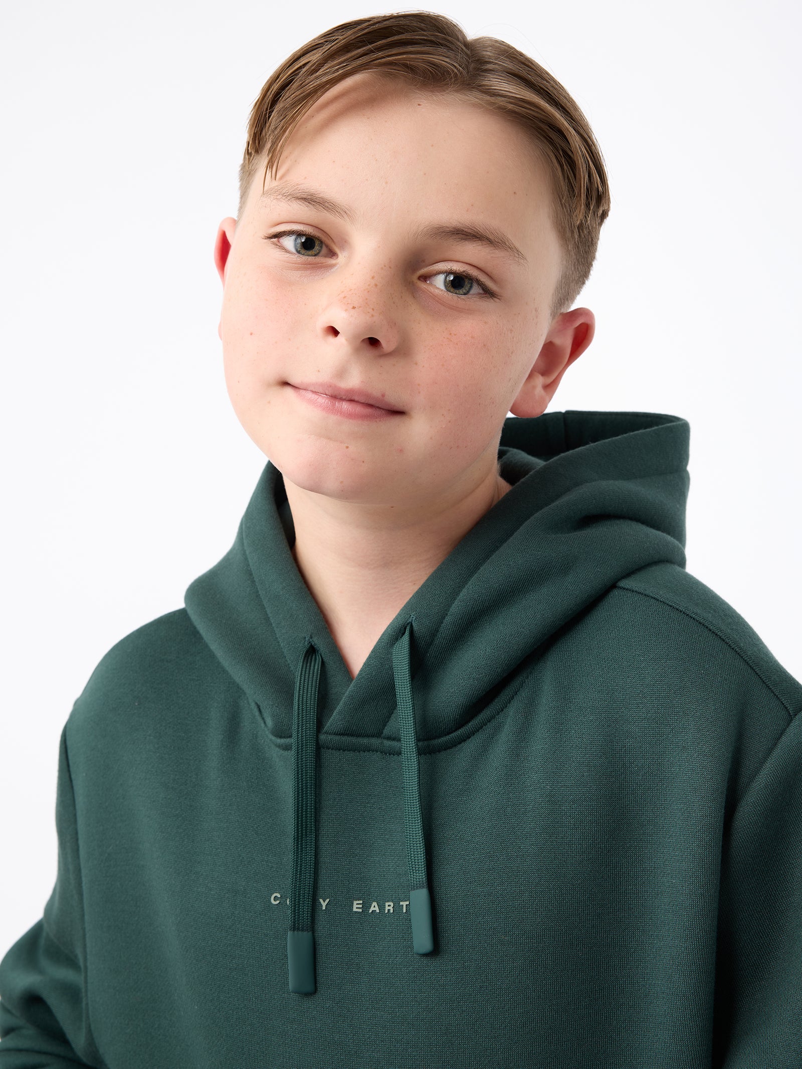 A young individual with short hair beams at the camera, dressed in a dark green Kid's CityScape Hoodie from Cozy Earth. The plain white background accentuates their presence. 