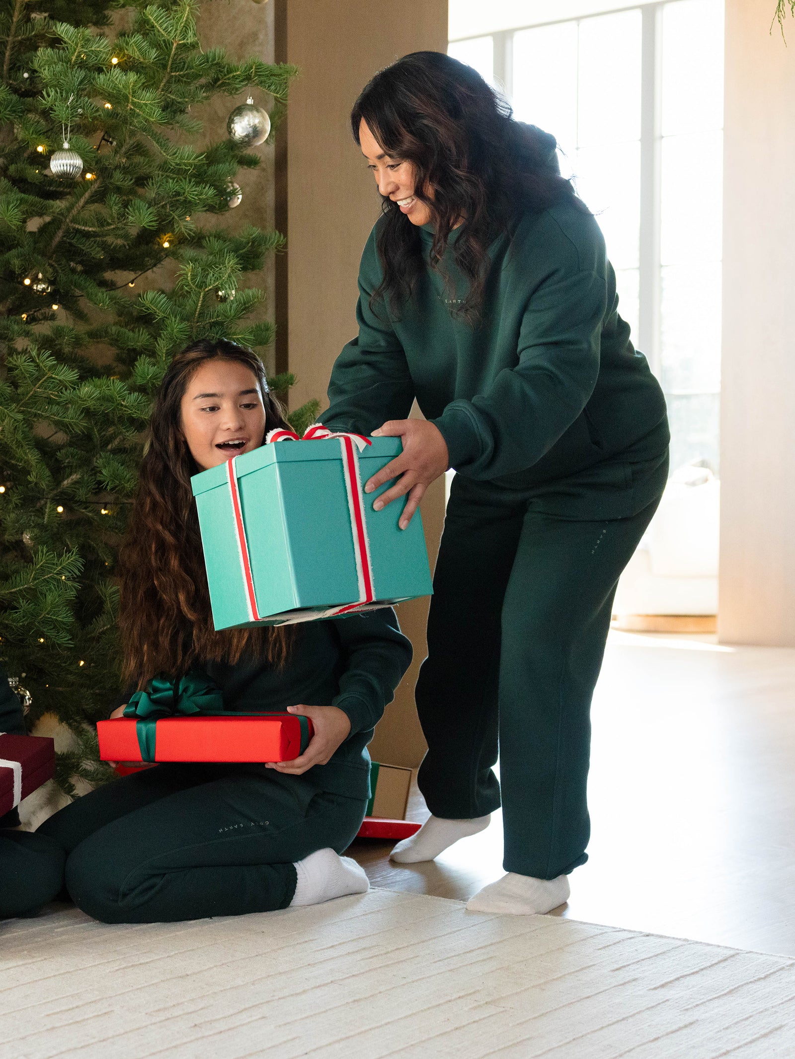 A woman, dressed in the Women's CityScape Sweatpant by Cozy Earth, hands a wrapped gift to a surprised-looking girl near a Christmas tree. Both are wearing matching green outfits from Cozy Earth, and the scene is filled with holiday decorations and soft natural light. 