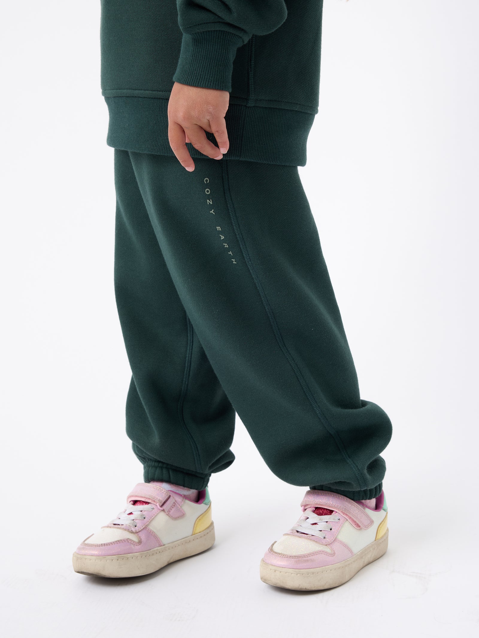 A person is wearing dark green Kid's CityScape Sweatpants from Cozy Earth, paired with a matching sweater. They are also sporting pink and white sneakers, with a plain white background. 