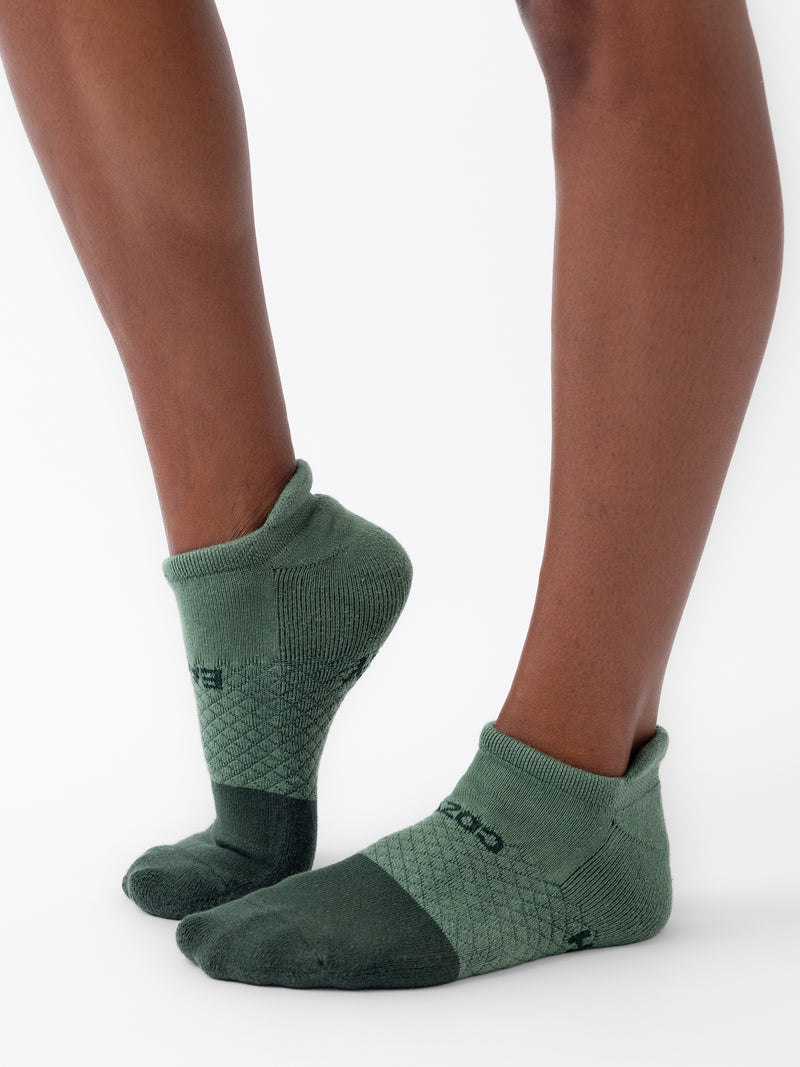 Close-up of a pair of feet wearing socks from the Cozy Earth Essential Ankle Sock 2-Pack. The green ankle socks feature a textured pattern and a darker green color on the toes and heel. The plain white background highlights the socks and legs perfectly.