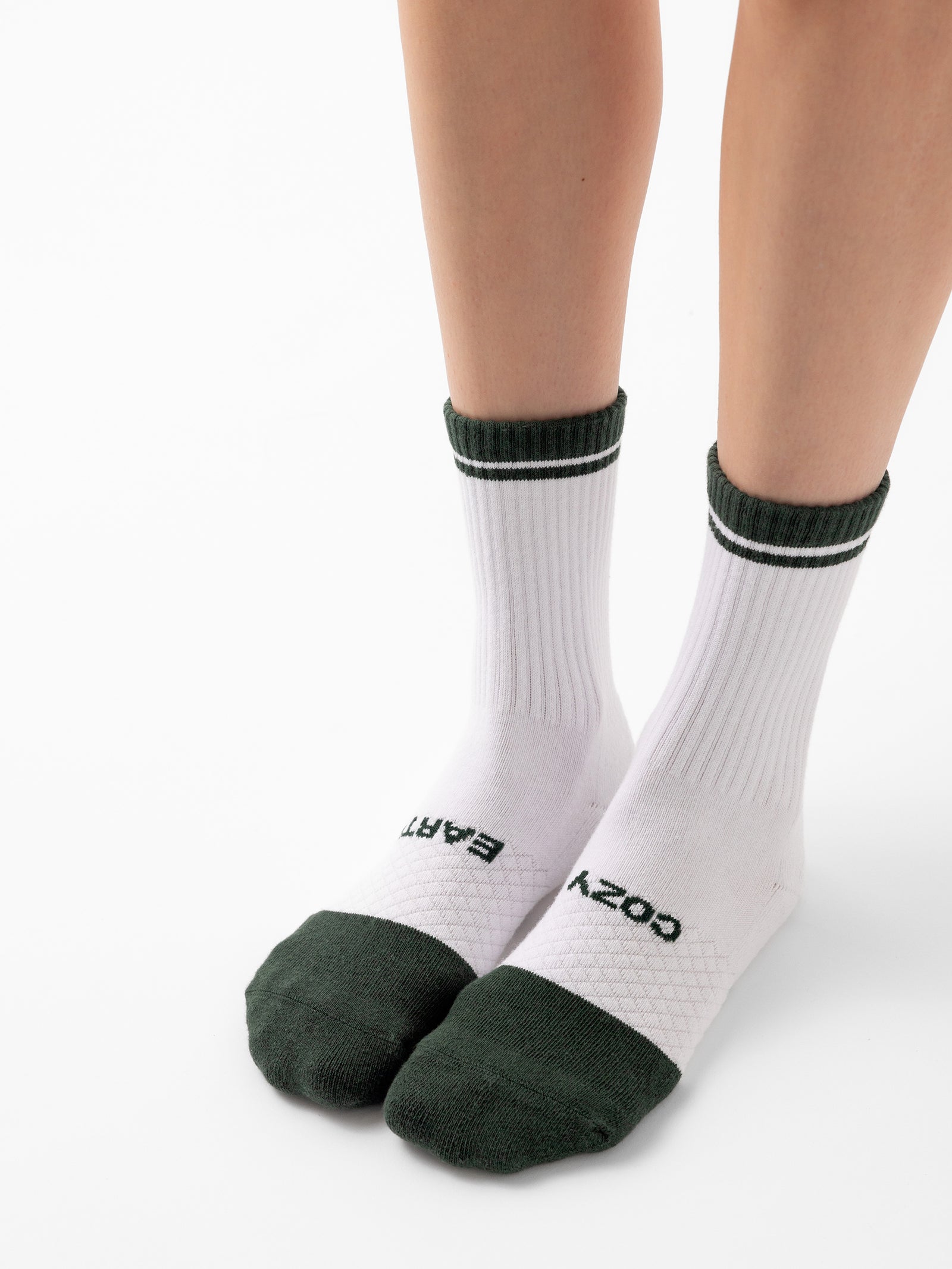 A person is standing on a white surface wearing socks from the Essential Calf Sock 2-Pack by Cozy Earth. The socks feature white bodies with green toes, heels, and top bands. The left sock displays the word "COZY" in black text, while the right sock shows "EARTH. 