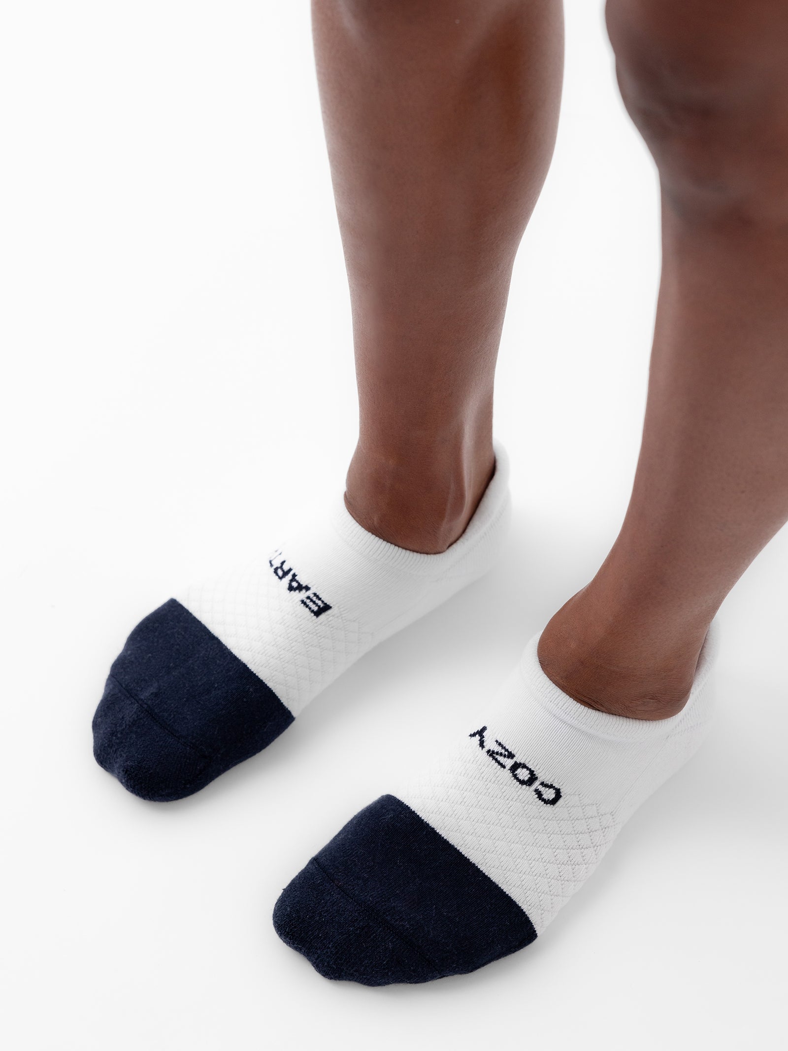 A person is standing on a white surface wearing white ankle socks with black toes from the Essential Ankle Sock 4-Pack by Cozy Earth. One sock has the word "EARTH" labeled on the top, and the other sock has "COZY" labeled on it. 