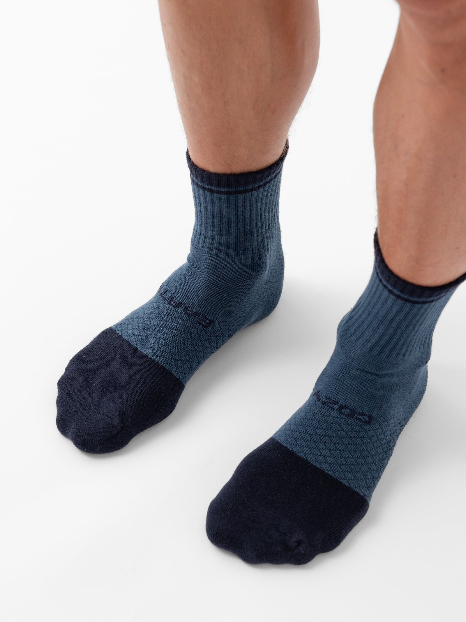 Close-up of a person wearing Cozy Earth's Essential Quarter Socks from the 4-Pack collection. The socks feature a textured pattern, slight ribbing near the ankle, and dark blue toes and heels. The person is standing on a white surface with their calves visible above the blue crew socks. 