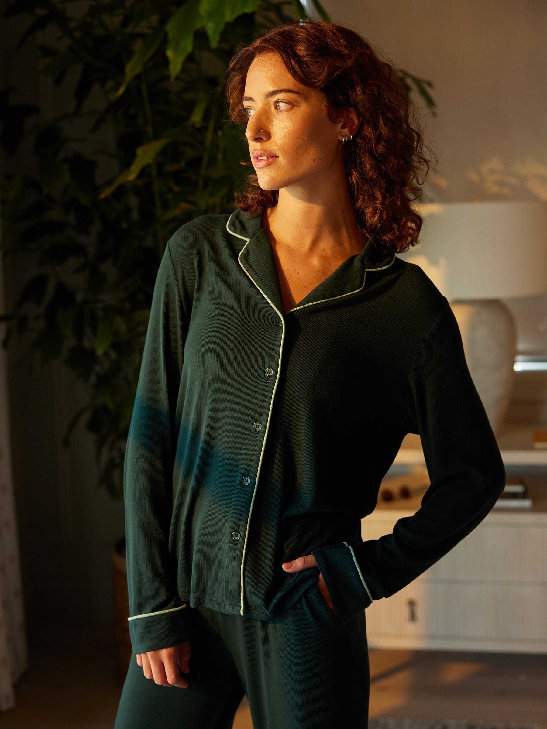 A person with curly hair stands indoors, dressed in a Cozy Earth Women's Bamboo Rib Knit Classic Long Sleeve Pajama Top in dark green with white piping. Their right hand rests in a pocket as they look pensively to the side. A potted plant and furniture are visible in the softly lit background. 