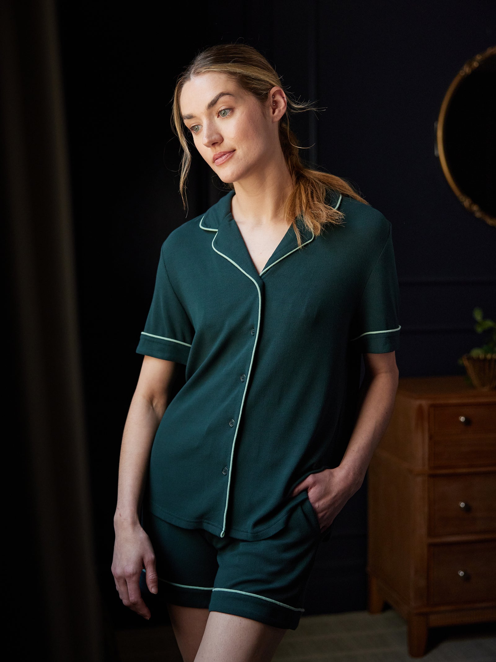 A person with long hair stands by a dark-colored wall wearing the Cozy Earth Women's Bamboo Rib Knit Classic Short Sleeve Pajama Top in dark green with white piping. One hand is in their pocket while the other rests relaxed by their side. A wooden dresser and a small potted plant are visible in the background. 