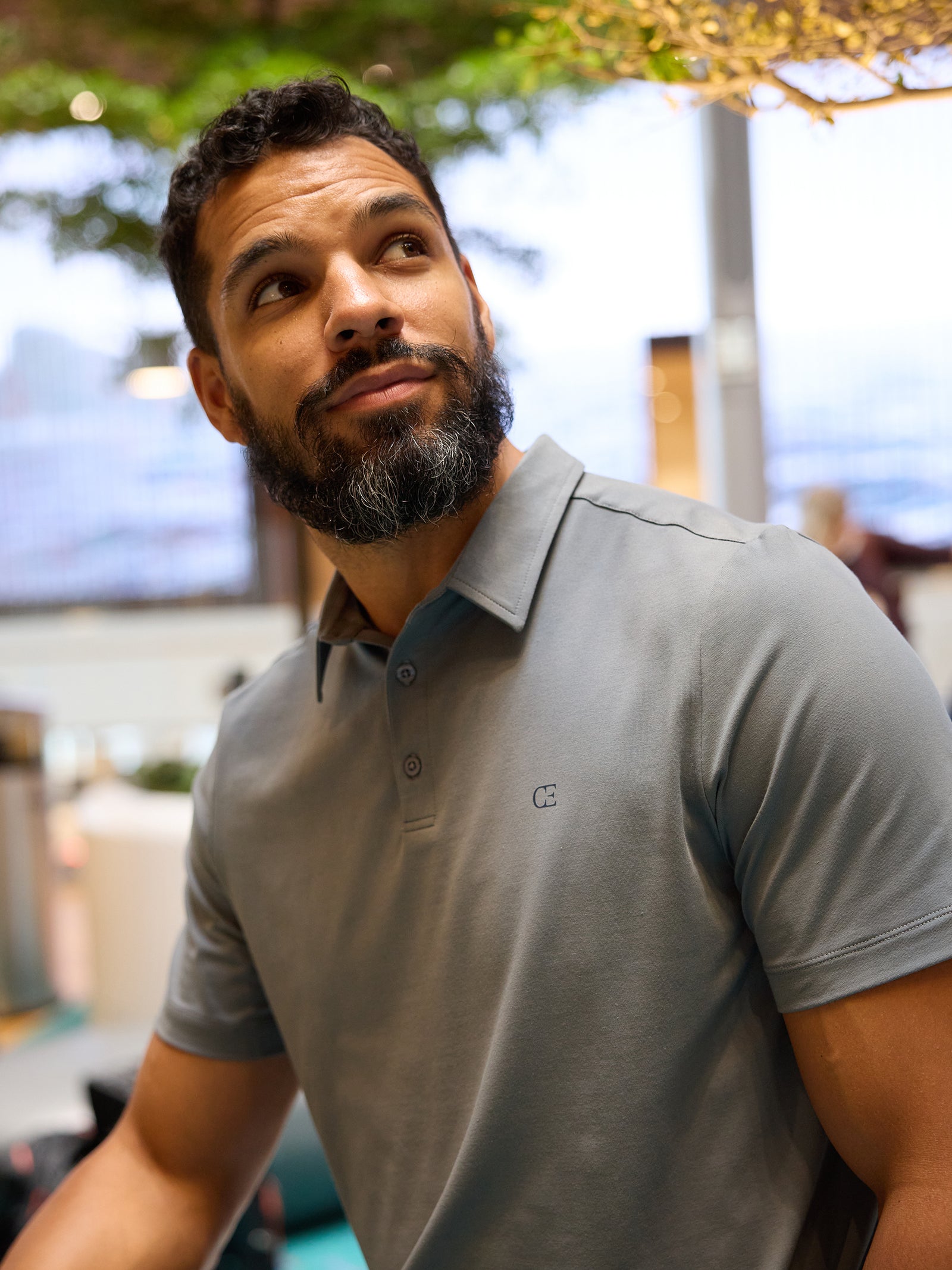 A man with a beard and mustache is wearing the Men's Everyday Polo from Cozy Earth in gray, looking upwards. The background features a blurred indoor setting with some greenery and soft lighting. 