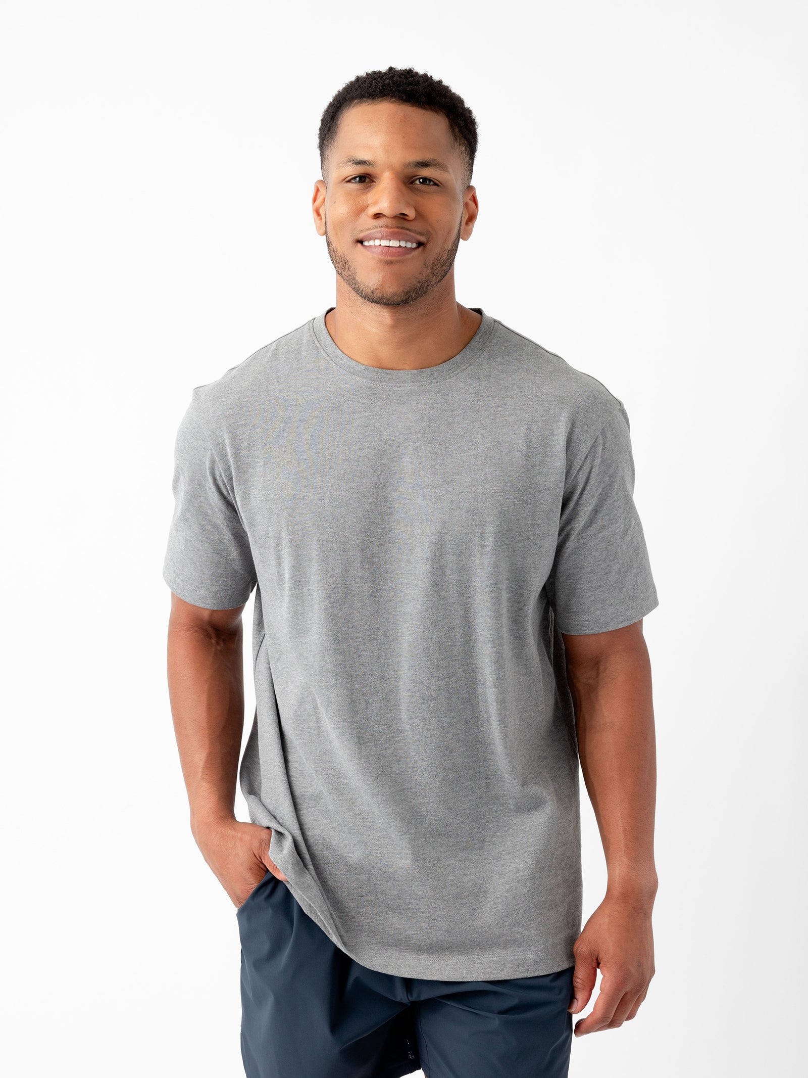 A smiling person with short hair, wearing a Cozy Earth Men's All Day Tee in gray and dark blue pants, stands against a plain white background. One hand is placed in their pants pocket, conveying an overall casual and relaxed appearance. 