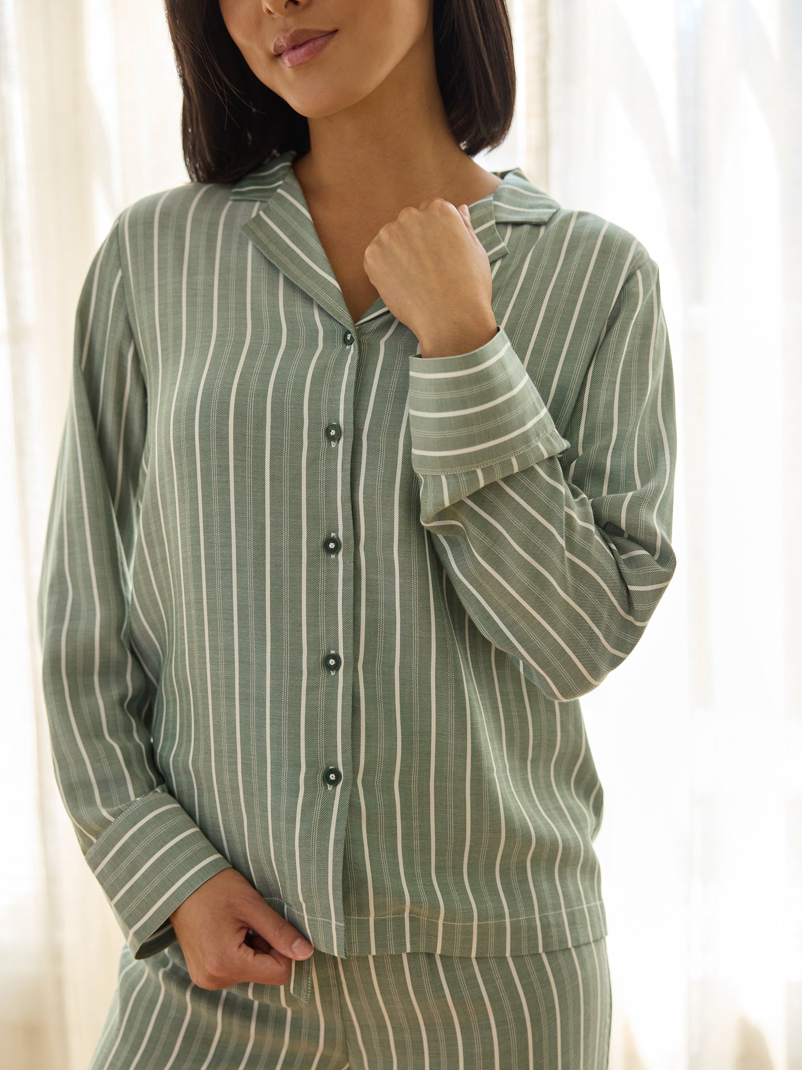 A person wearing the Women's Soft Woven Long Sleeve Pajama Top by Cozy Earth stands in a softly lit room. The green and white striped pajama top features buttons down the front and a collar, which the person is gently holding with one hand. 