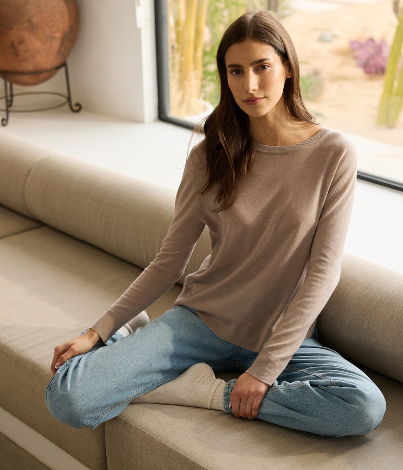 A woman with long brown hair sits cross-legged on a beige couch near a window, wearing the Women's AirKnit Crewneck Sweater by Cozy Earth and blue jeans. They have a calm expression, and the background features a round pottery piece and a garden view. 