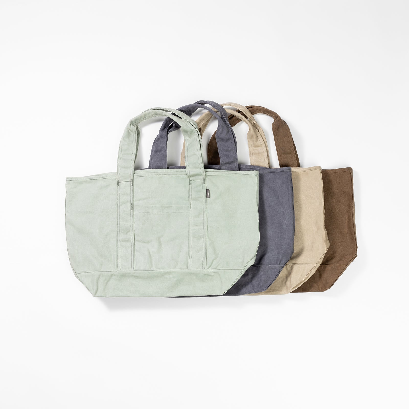 Five Waxed Canvas Tote bags from Cozy Earth are displayed in a row against a white background. The bags are arranged in descending order from left to right by color: light green, dark gray, light gray, beige, and brown. Each bag features two handles and a front pocket. 