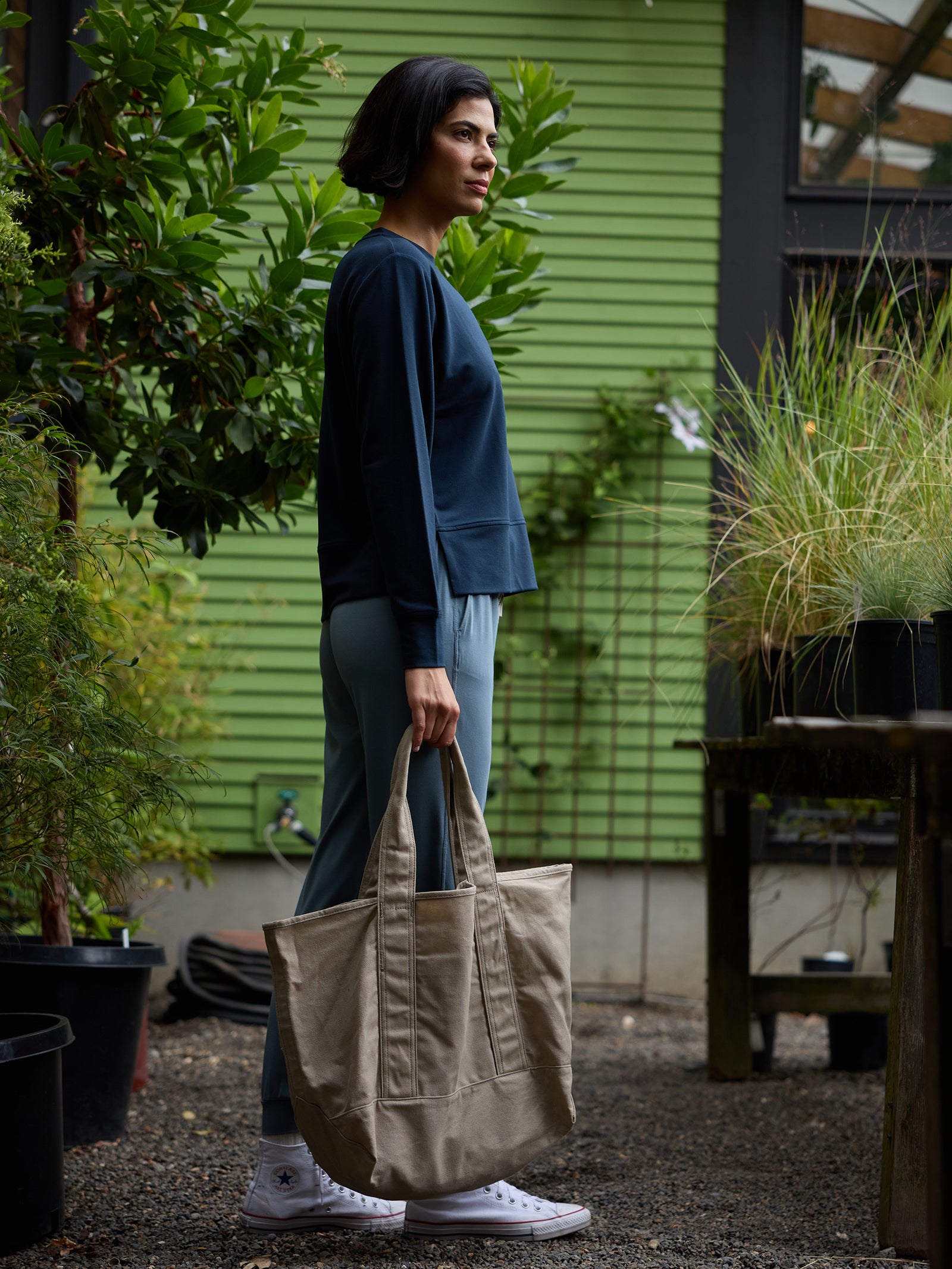 A person in a navy sweater and light blue pants stands in a garden area holding a large Waxed Canvas Tote by Cozy Earth. They have short dark hair and are wearing white sneakers. The background features green plants, trees, and a green building with horizontal siding. 